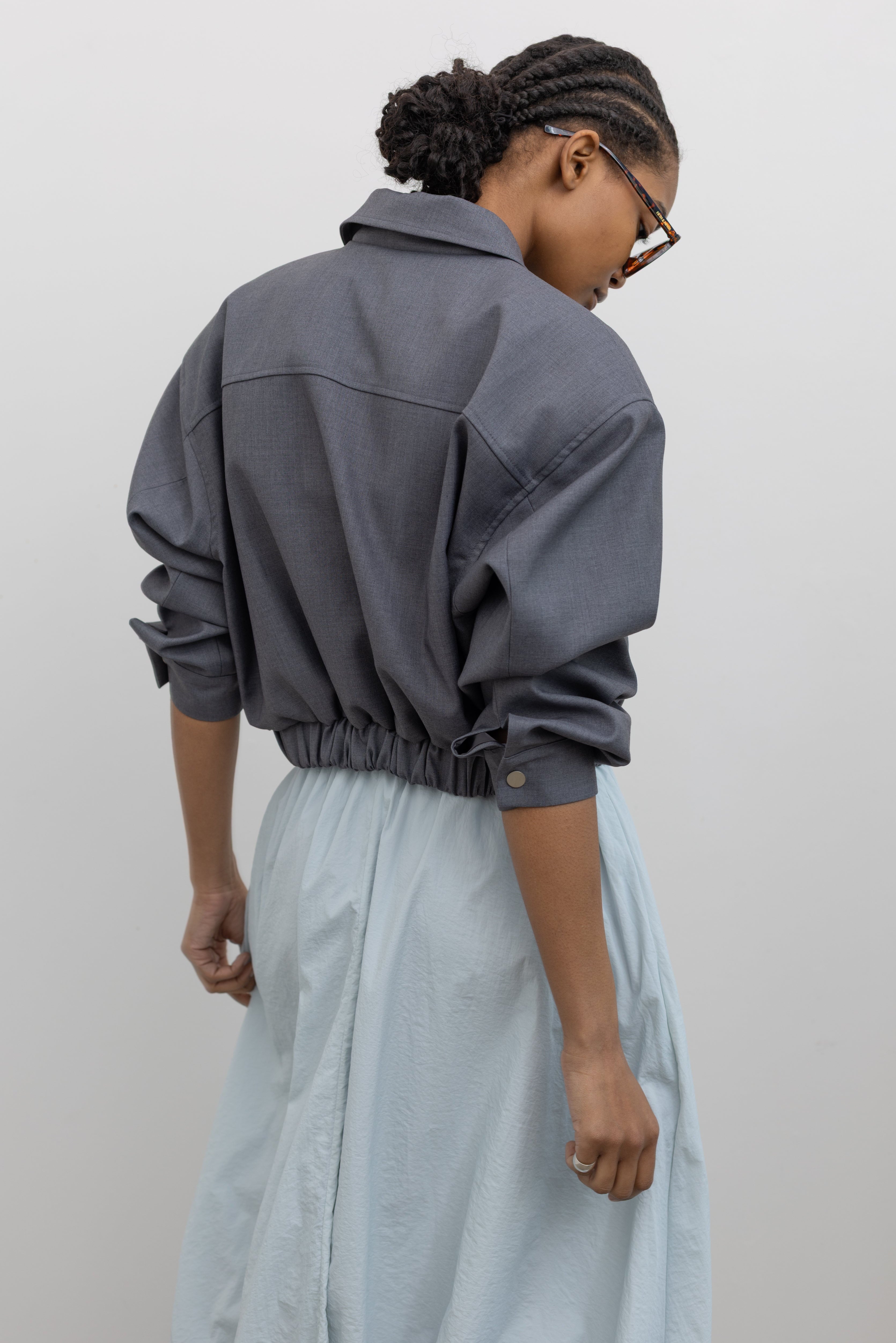 Back view of a woman wearing chic and refined spring fashion in an Alas Eius label charcoal cropped tailored zip jacket featuring an elastic hem for a flattering tailored look at an Alas Eius store. 