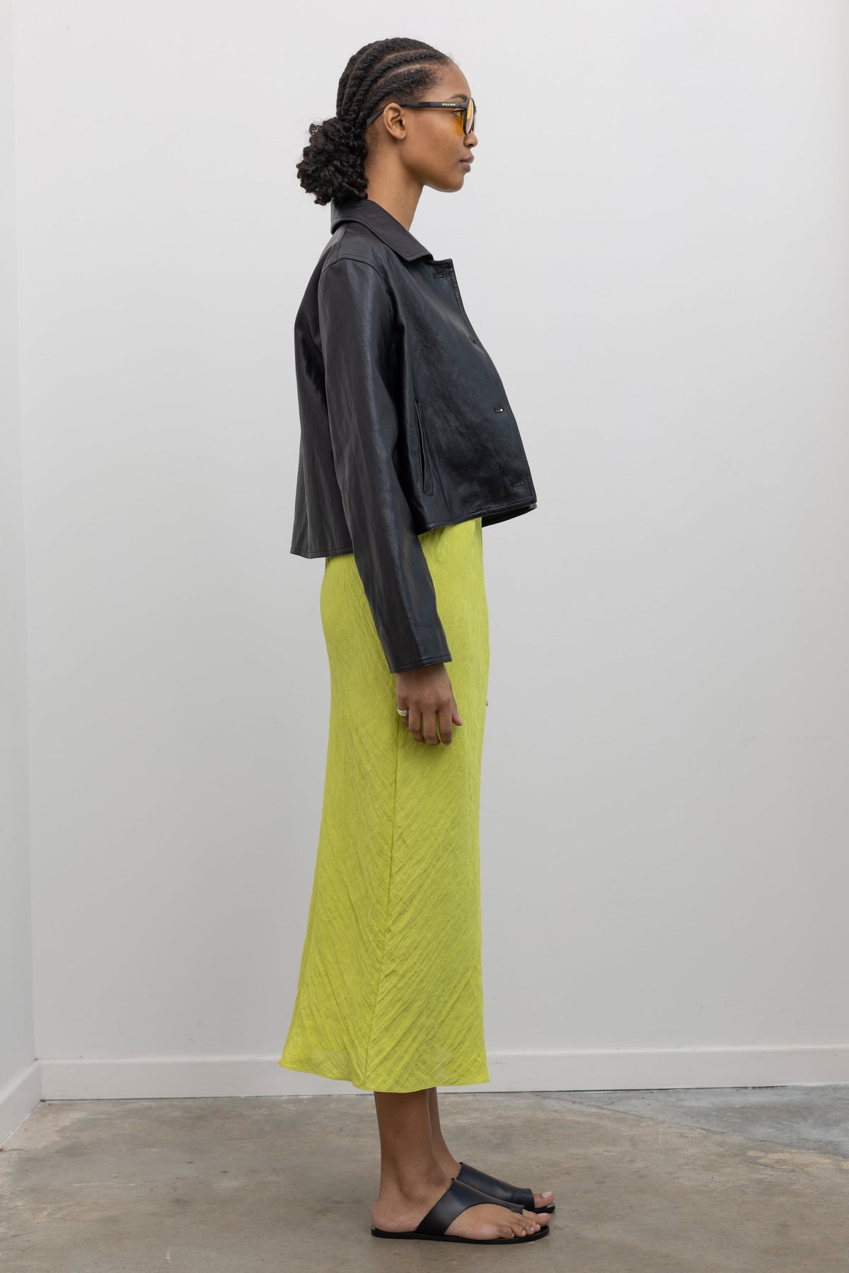 Full length side view of a woman posing wearing chic and refined Alas Eius Label Black Cropped Vegan Leather Collared Jacket with a chartreuse midi dress and ATP black leather cutout sandals at Alas Eius.  