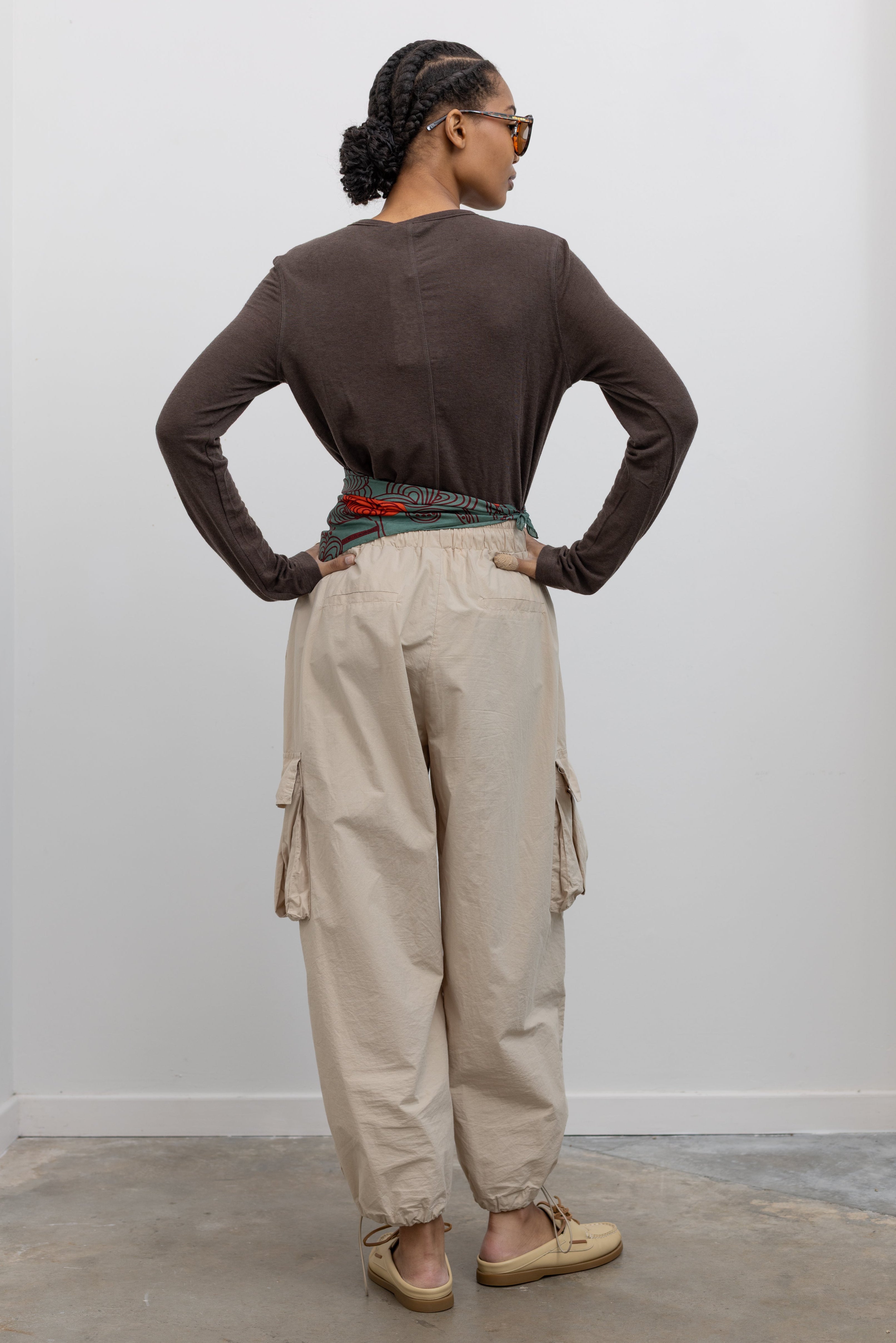 Back view of Woman standing wearing modern fashion in a brown long-sleeved top with a pair of Alas Eius Label Tan Wide Leg Cargo Pants with leather flat mule shoes and printed scarf around waist at Alas Eius