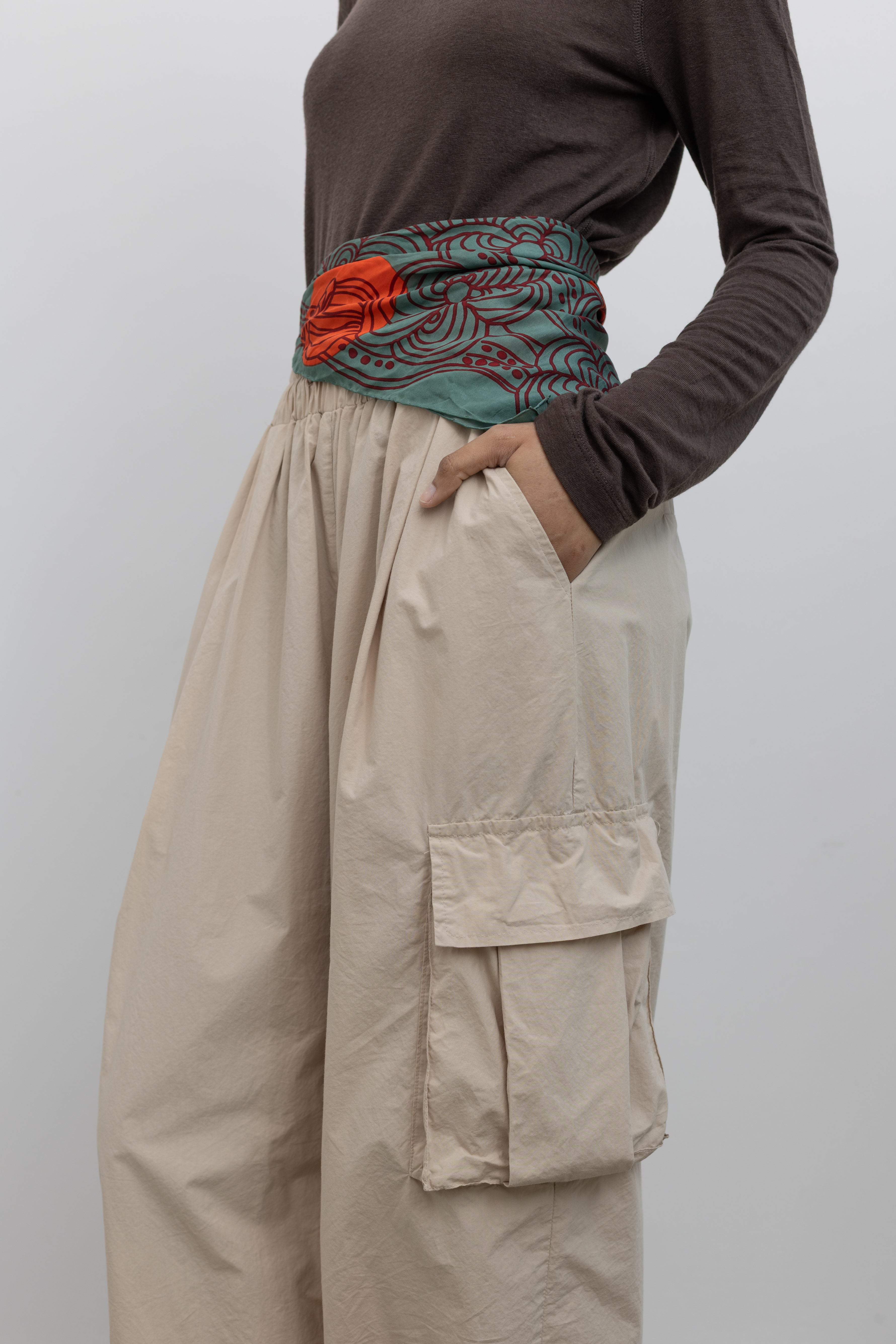 Woman standing wearing modern fashion in a brown long-sleeved top with a pair of Alas Eius Label Tan Wide Leg Cargo Pants with leather flat mule shoes and printed scarf around waist at Alas Eius