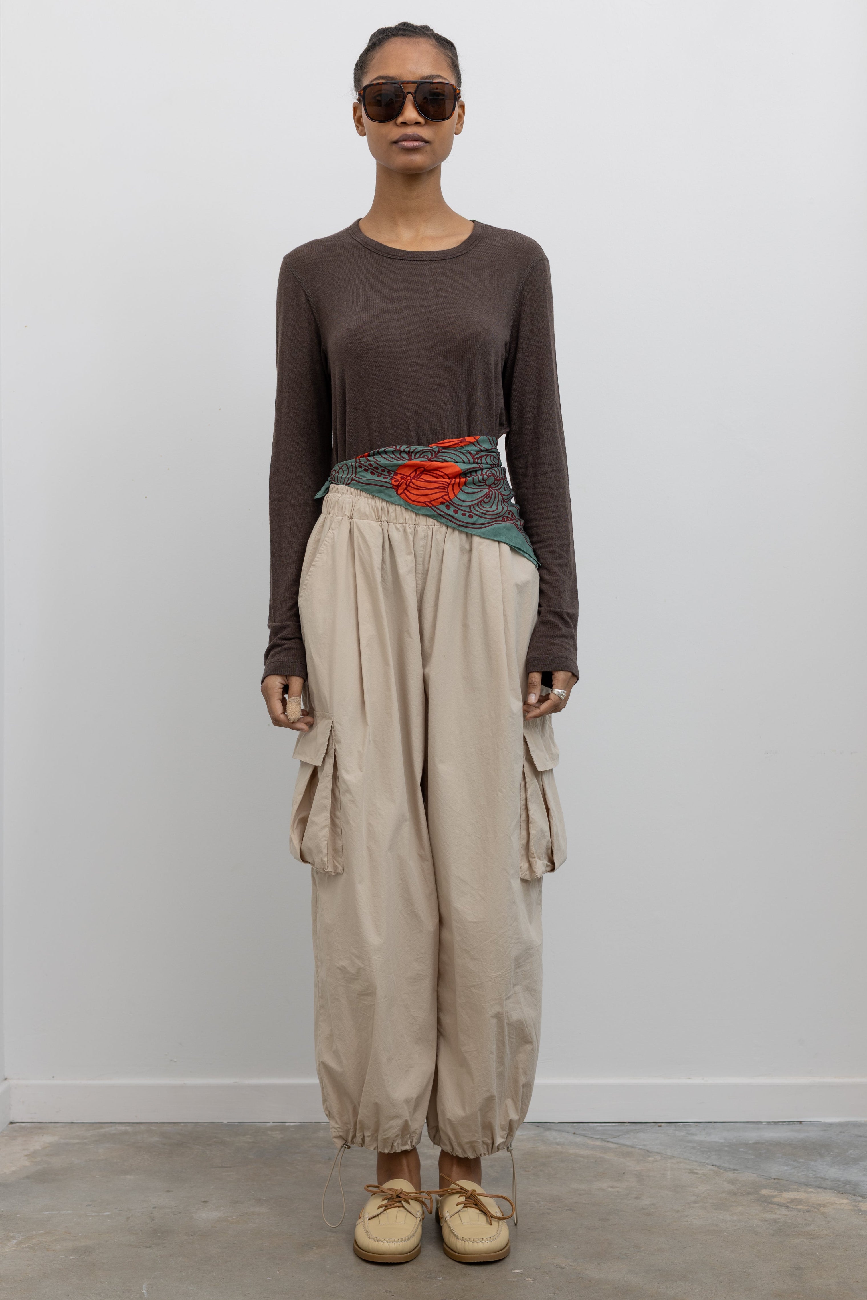 Front view of Woman standing wearing modern fashion in a brown long-sleeved top with a pair of Alas Eius Label Tan Wide Leg Cargo Pants with leather flat mule shoes and printed scarf around waist at Alas Eius