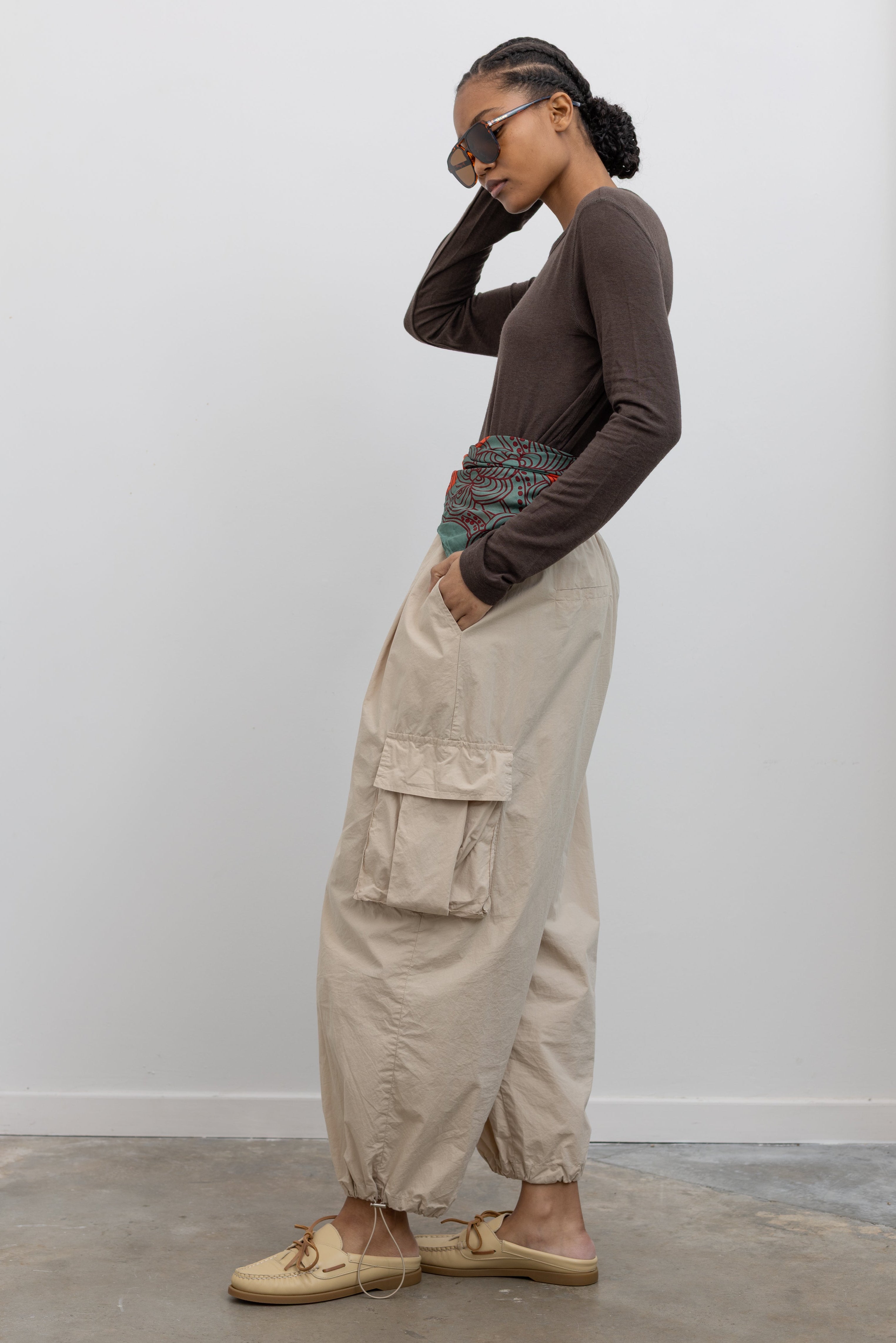 Side view of Woman standing wearing modern fashion in a brown long-sleeved top with a pair of Alas Eius Label Tan Wide Leg Cargo Pants with leather flat mule shoes and printed scarf around waist at Alas Eius