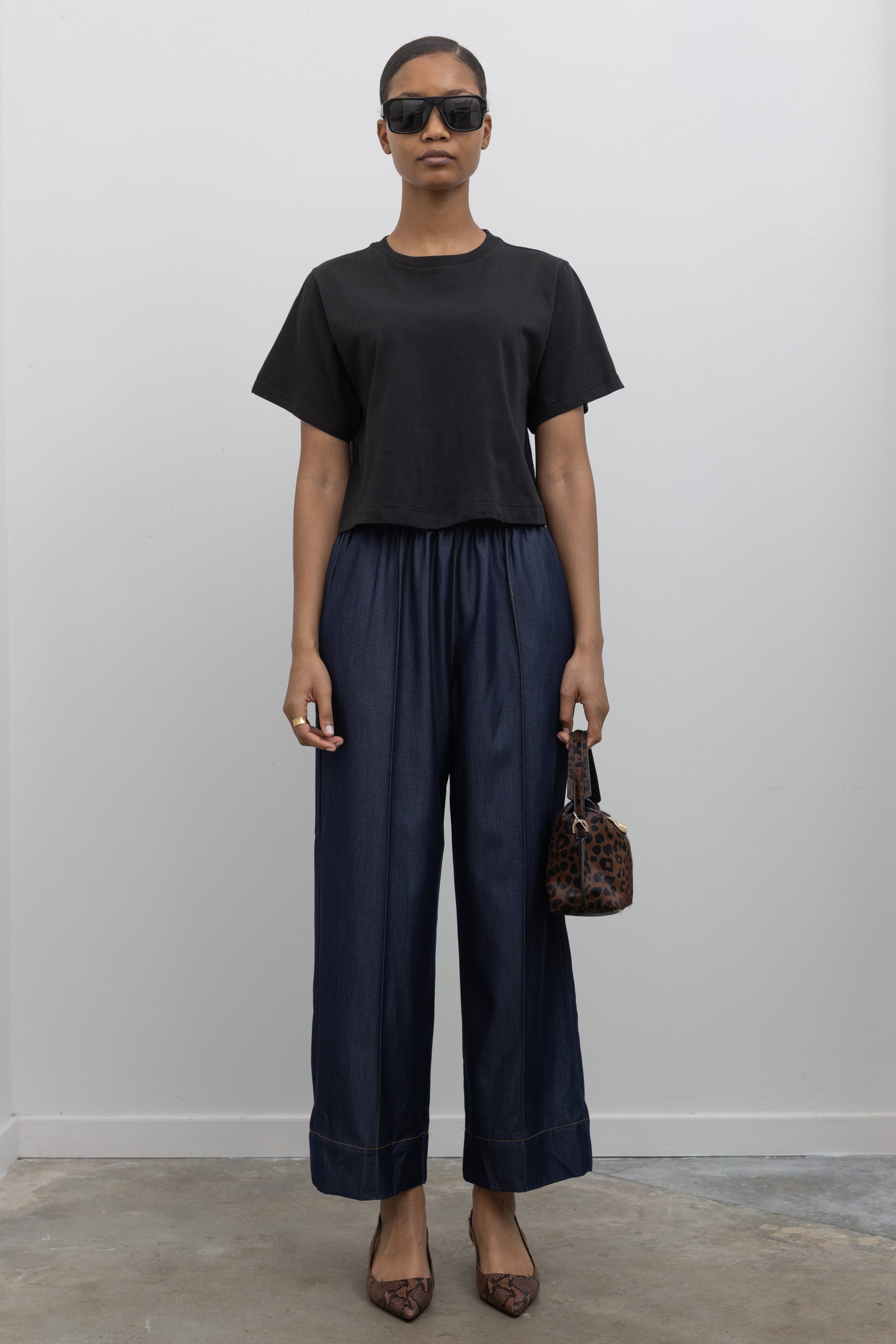 Front view of Woman standing wearing an everyday outfit with a black cotton short sleeved cropped t-shirt and Alas Eius Label Tencel Blend Dark Wash Tailored Wide Leg Pull On Pants with slingback kitten heels and accessories at Alas Eius. 