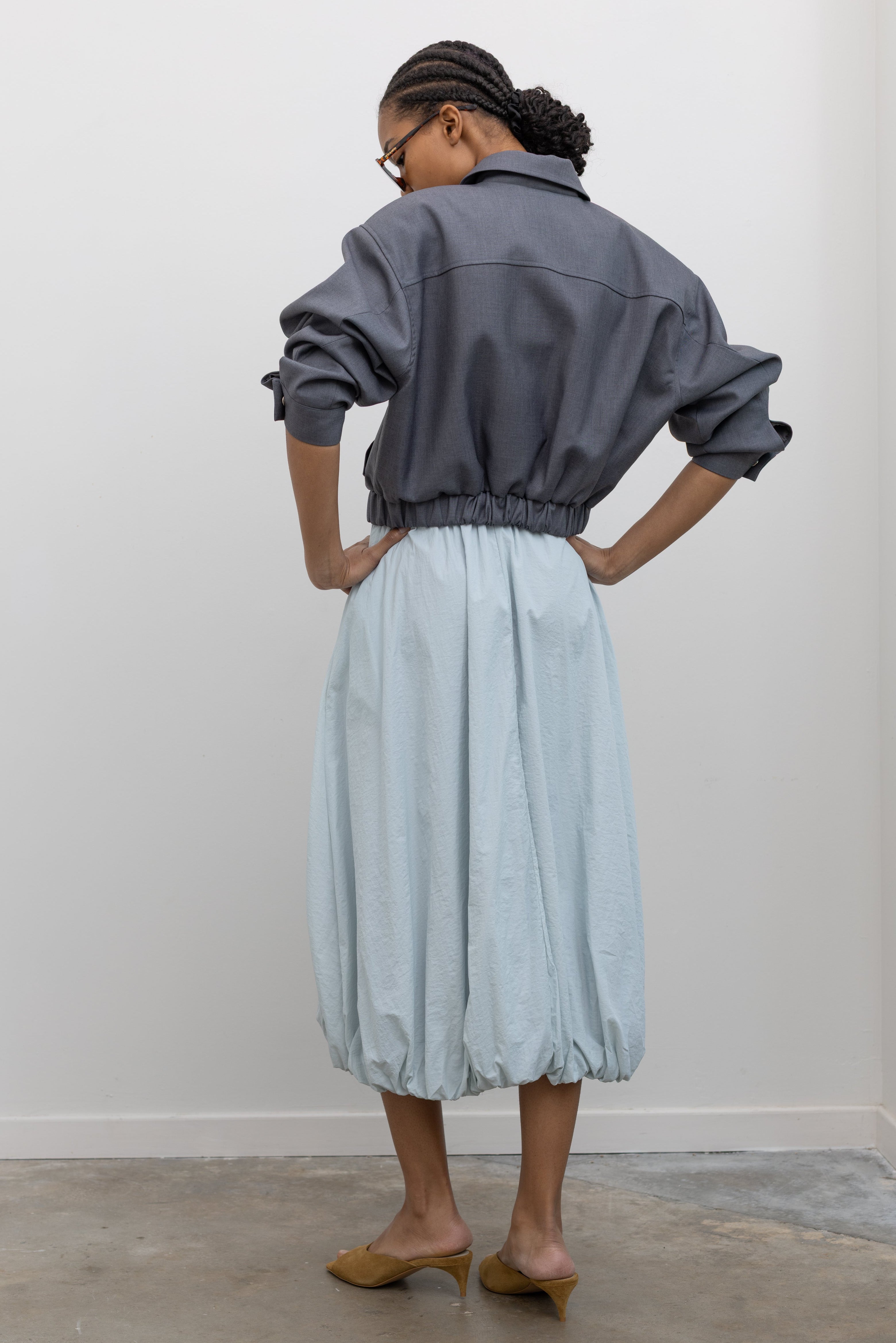 Back view of a woman wearing contemporary womenswear spring fashion in an Alas Eius label charcoal gray cropped tailored zip jacket featuring an elastic hem for a flattering cinched waist at an Alas Eius store. 