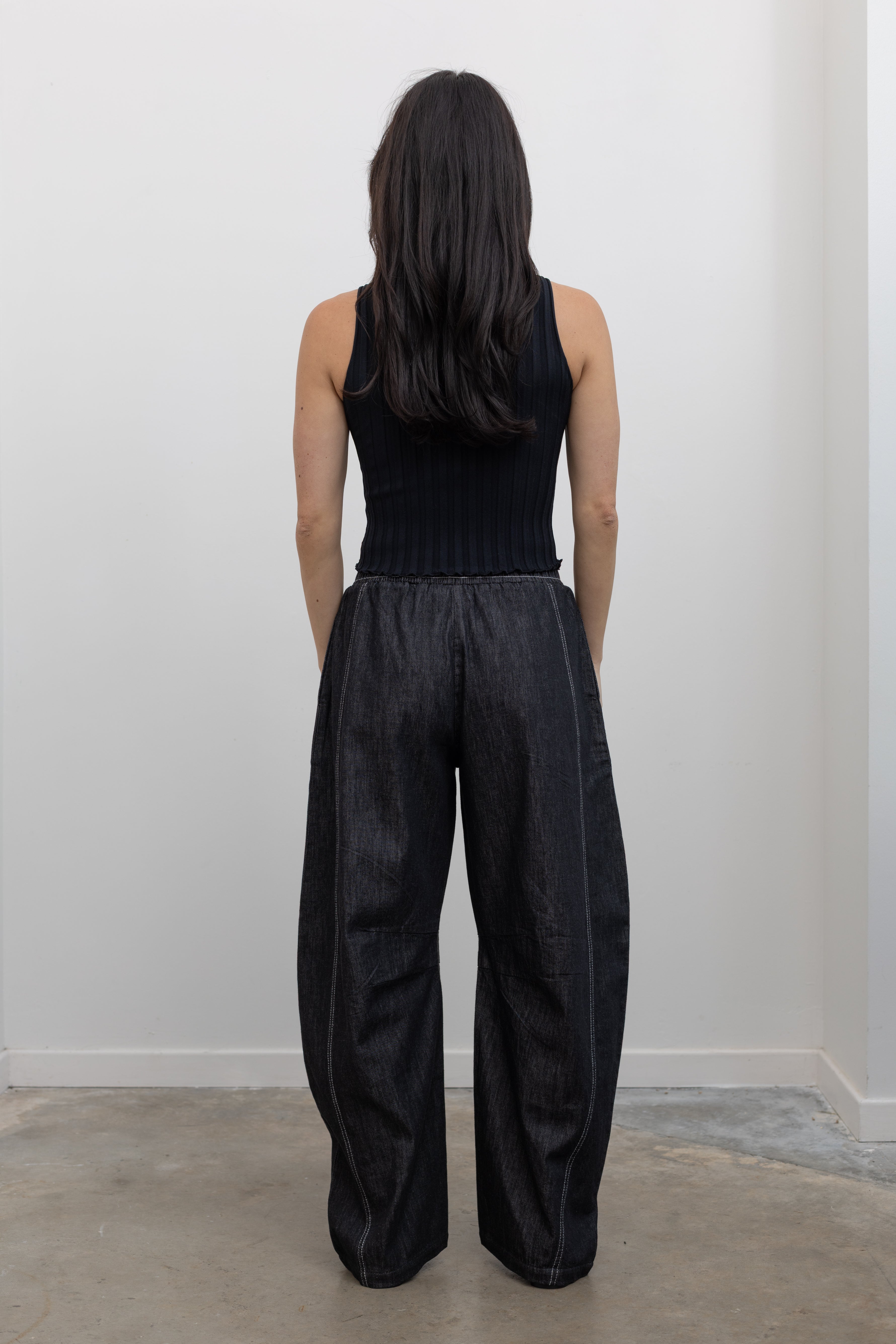 Full length back view of a woman standing wearing modern minimalist street wear in the Alas Eius label black cotton barrel leg pants featuring contrasting white double stitching, side seam pockets, and an elastic waistband paired with a black ribbed knit crop tank and ATP black leather cut out sandals at an Alas Eius store. 
