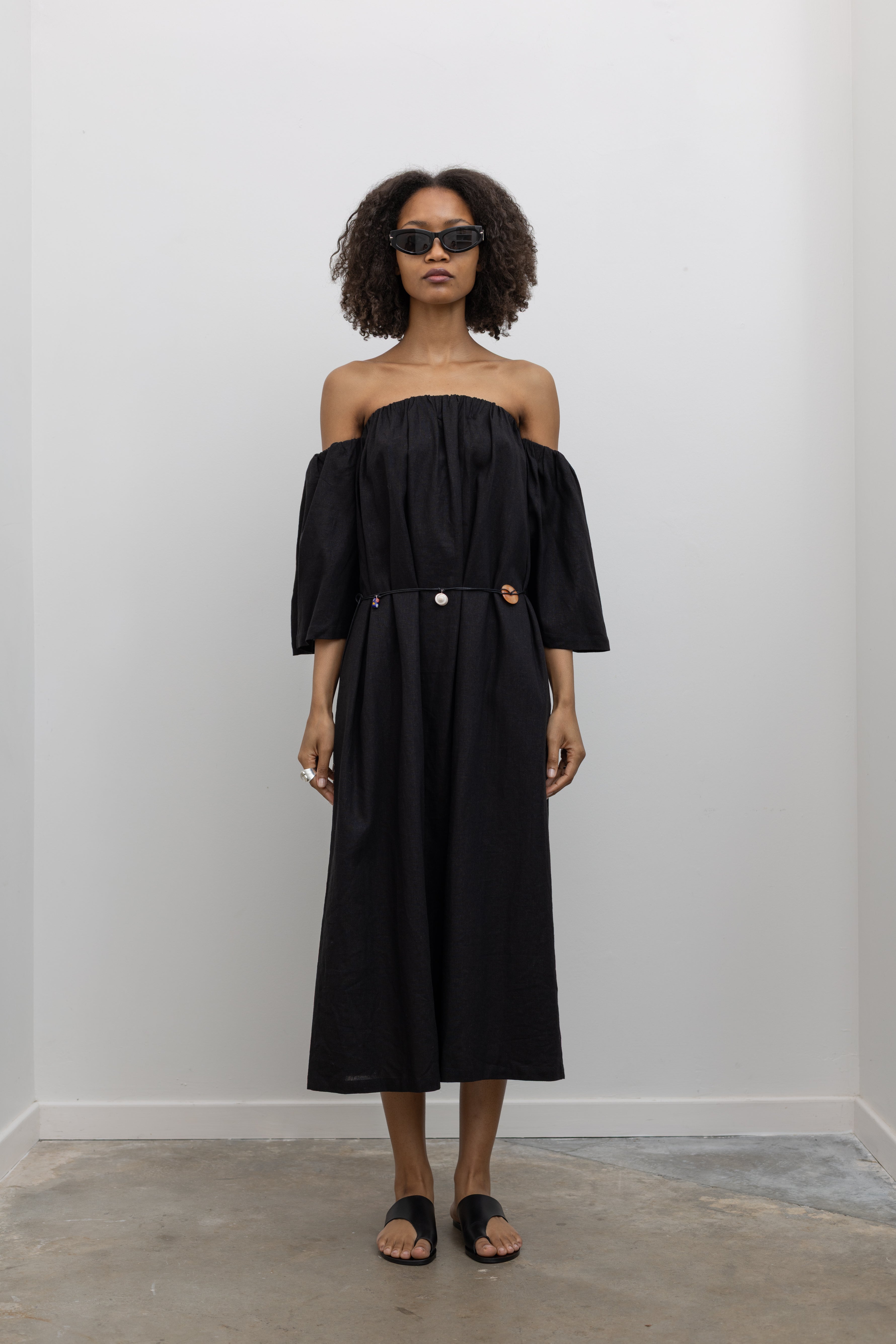 Front view of a woman standing wearing chic summer fashion in a Alas Eius label black linen midi dress featuring removable sleeves paired with a beaded belt at an Alas Eius store.