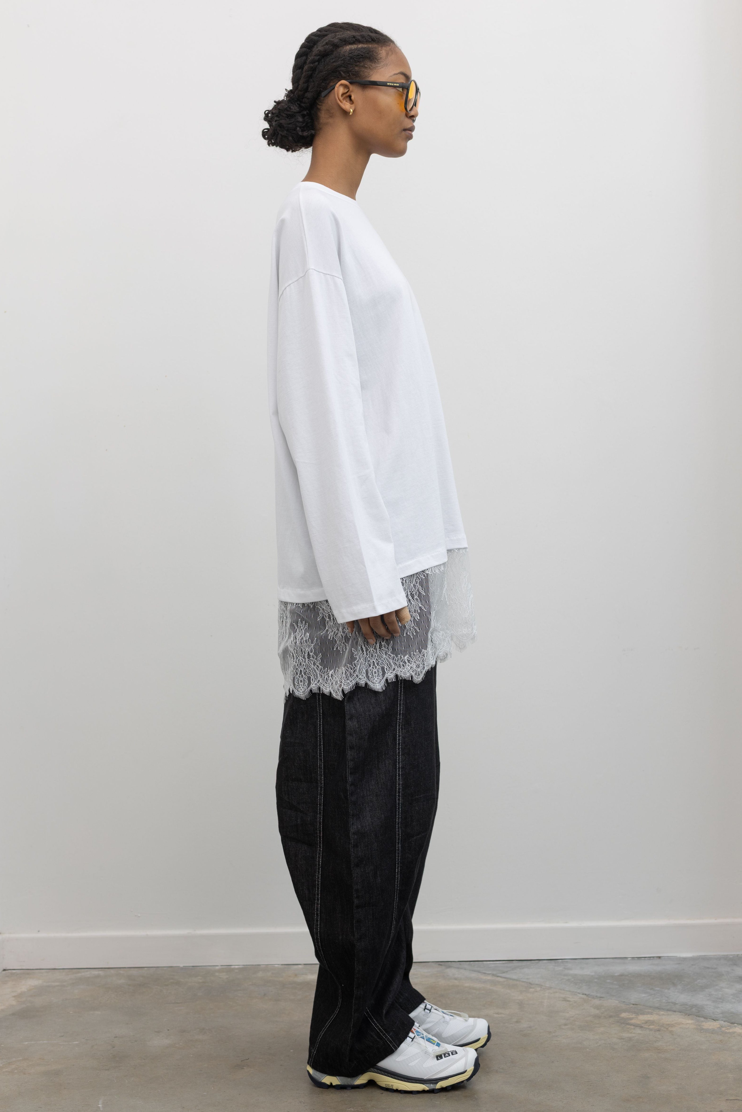 Full length side view of a woman standing wearing spring fashion in a 
versatile Alas Eius label white long-sleeve oversized tunic top with lace hemline at and Alas Eius store. 