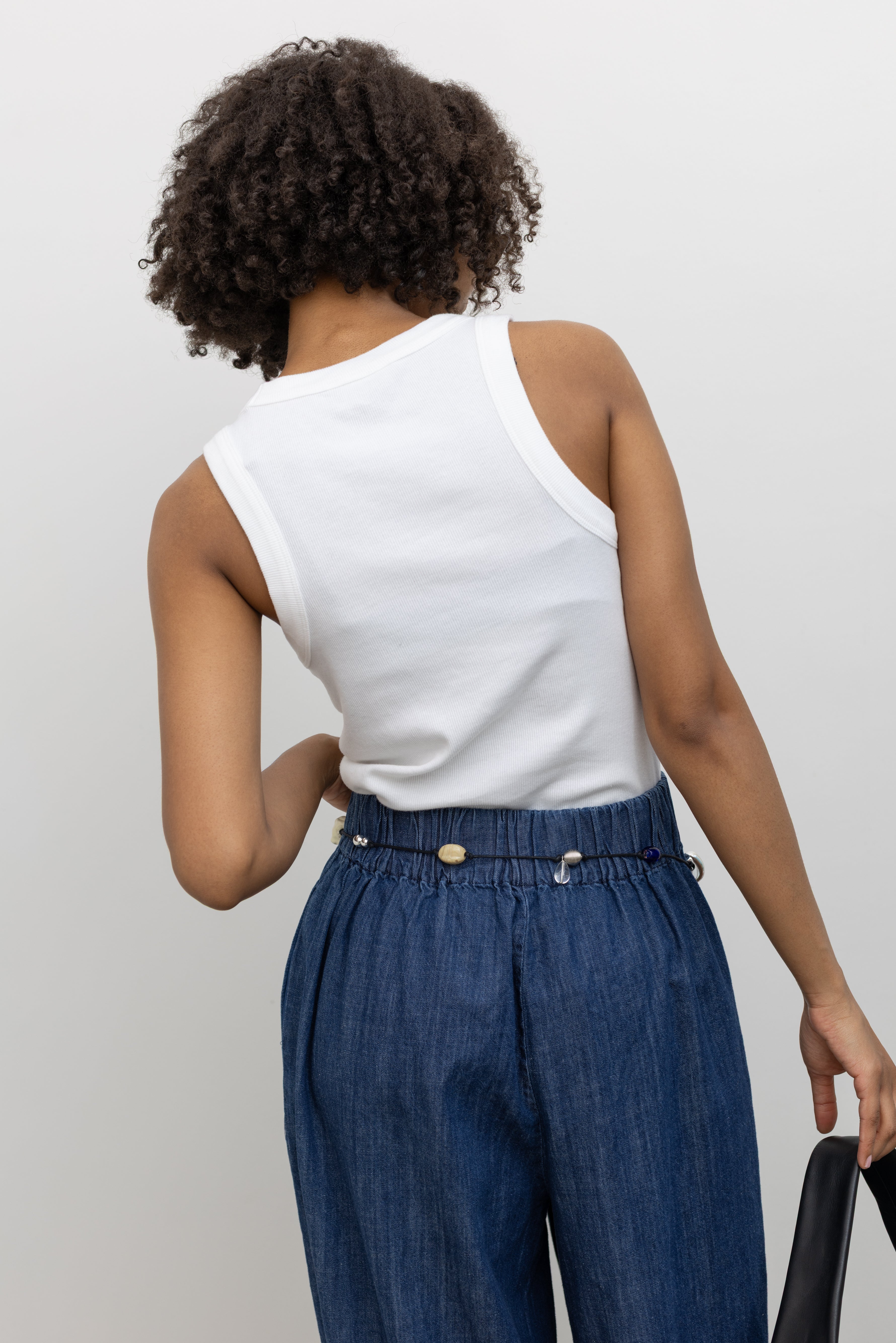 Model wearing an elevated basic in a Alas Eius label white cotton blend ribbed racerback tank top with contrasting embroidered AE logo on chest paired Collective blue cotton denim wide leg flowy pull on pant at Alas Eius. 