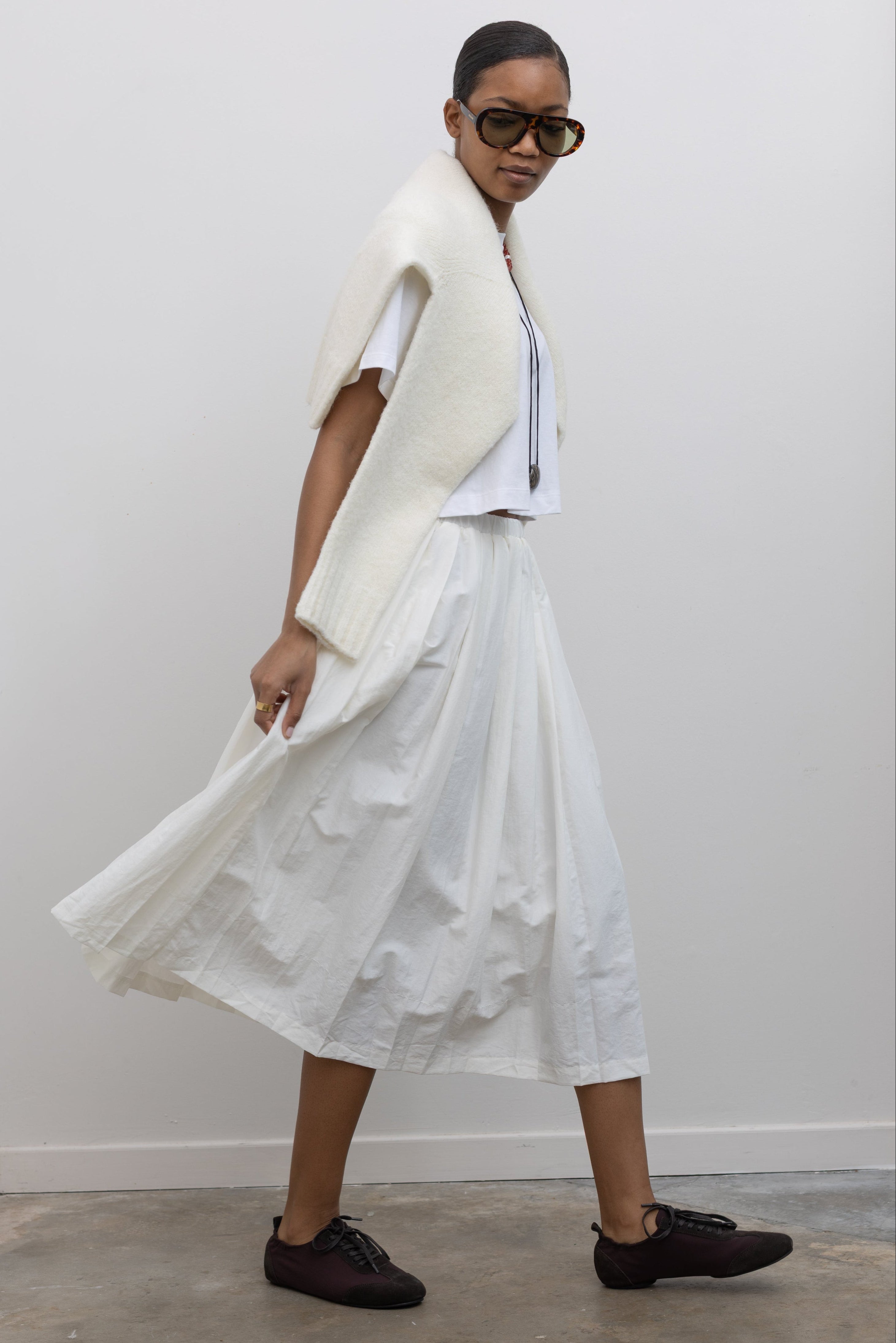 Woman in motion wearing capsule wardrobe essential style in an elegant Alas Eius Label white lightweight nylon pleated midi skirt, featuring a fully lined silhouette and finished with an elastic waistband and convenient side pockets. 