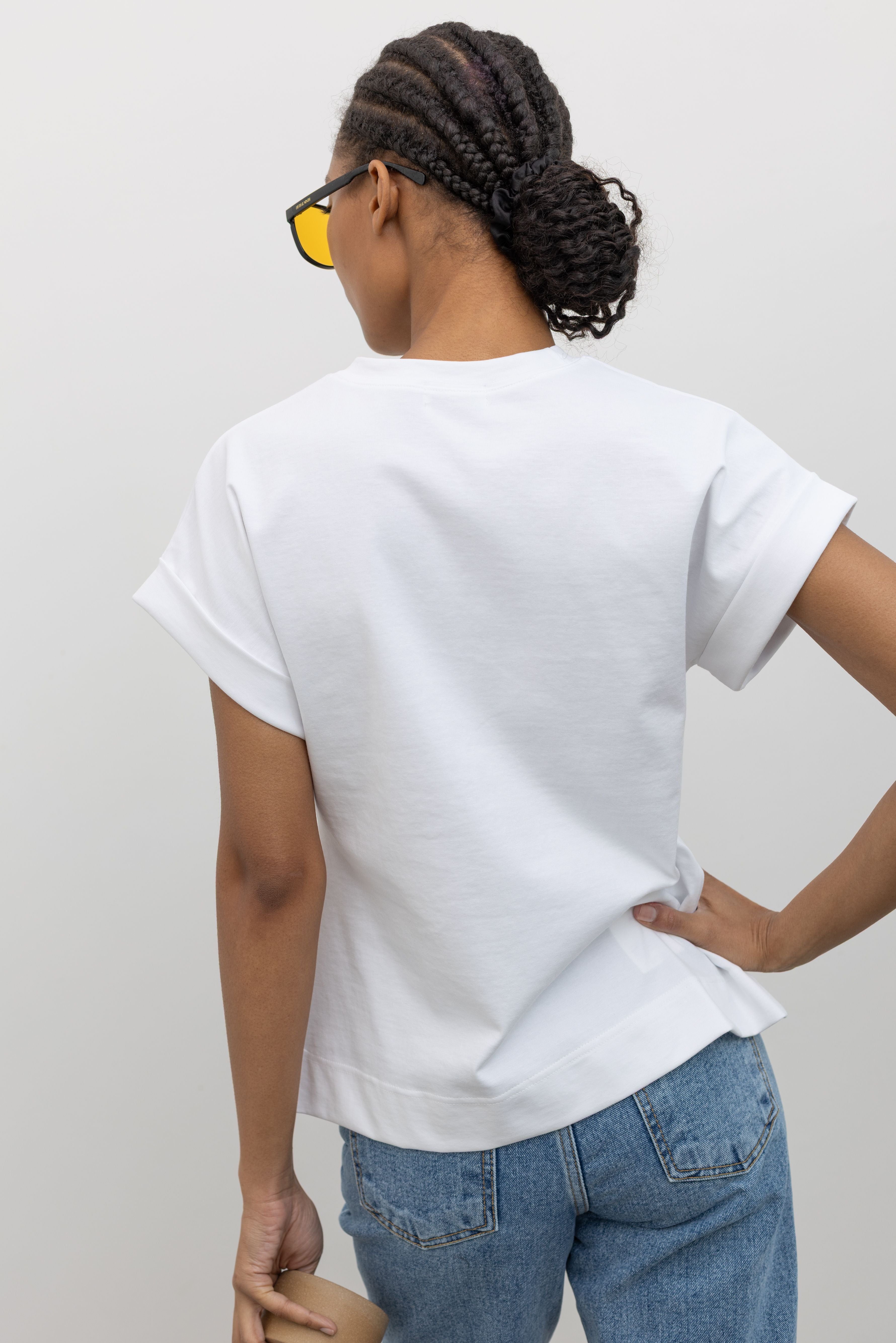 Back view of a woman posing with sunglasses wearing an elevated spring staple in a Alas Eius label white cinched waist rounded top designed stitched cuffed short sleeves, ruched side seams and an open v-cut vent at the front paired with light wash denim jeans at an Alas Eius store. 