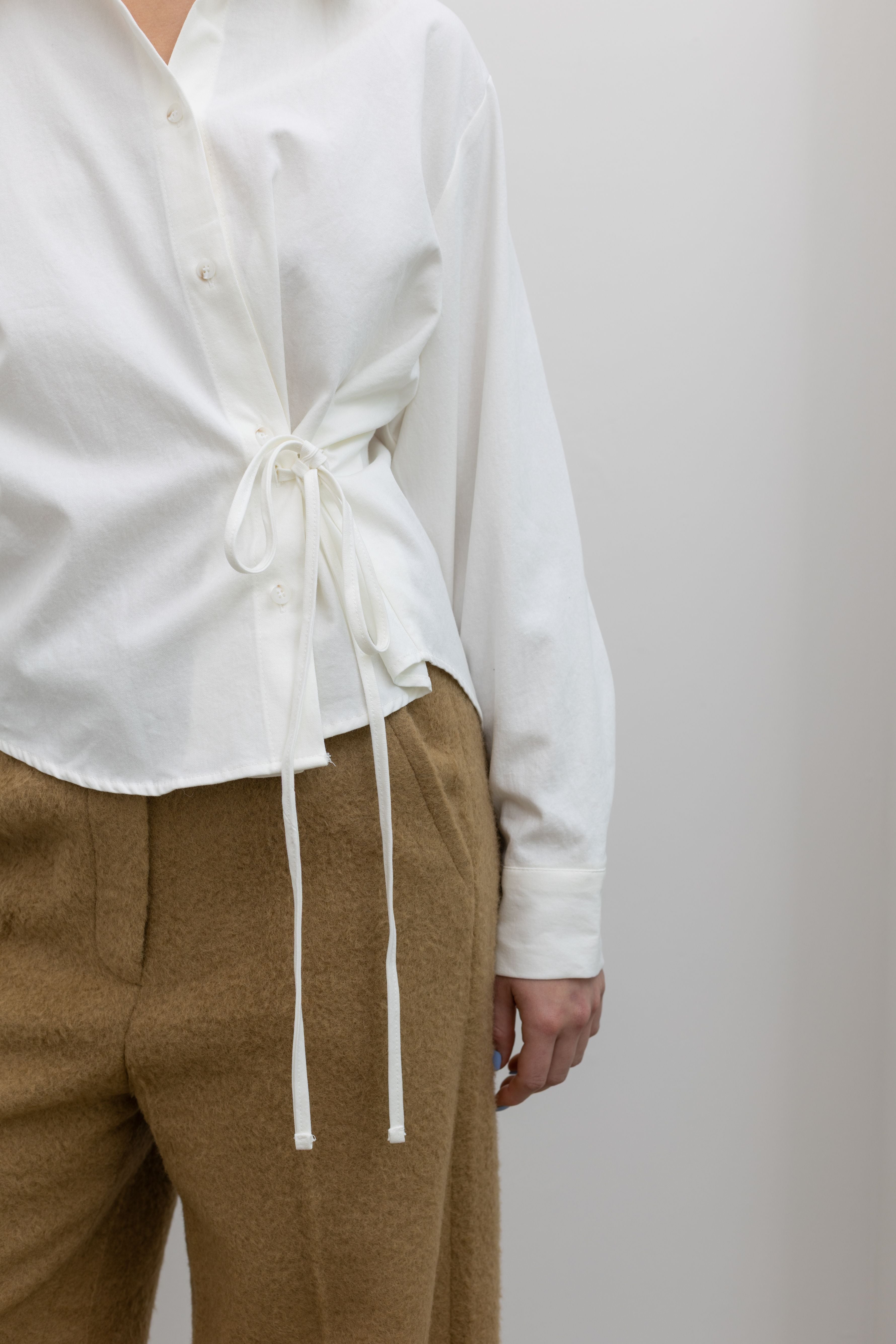 Close-up detailed view of a woman wearing an Alas Eius label white cotton side-cinched waist tie detail button down top paired with tan trousers at Alas Eius.