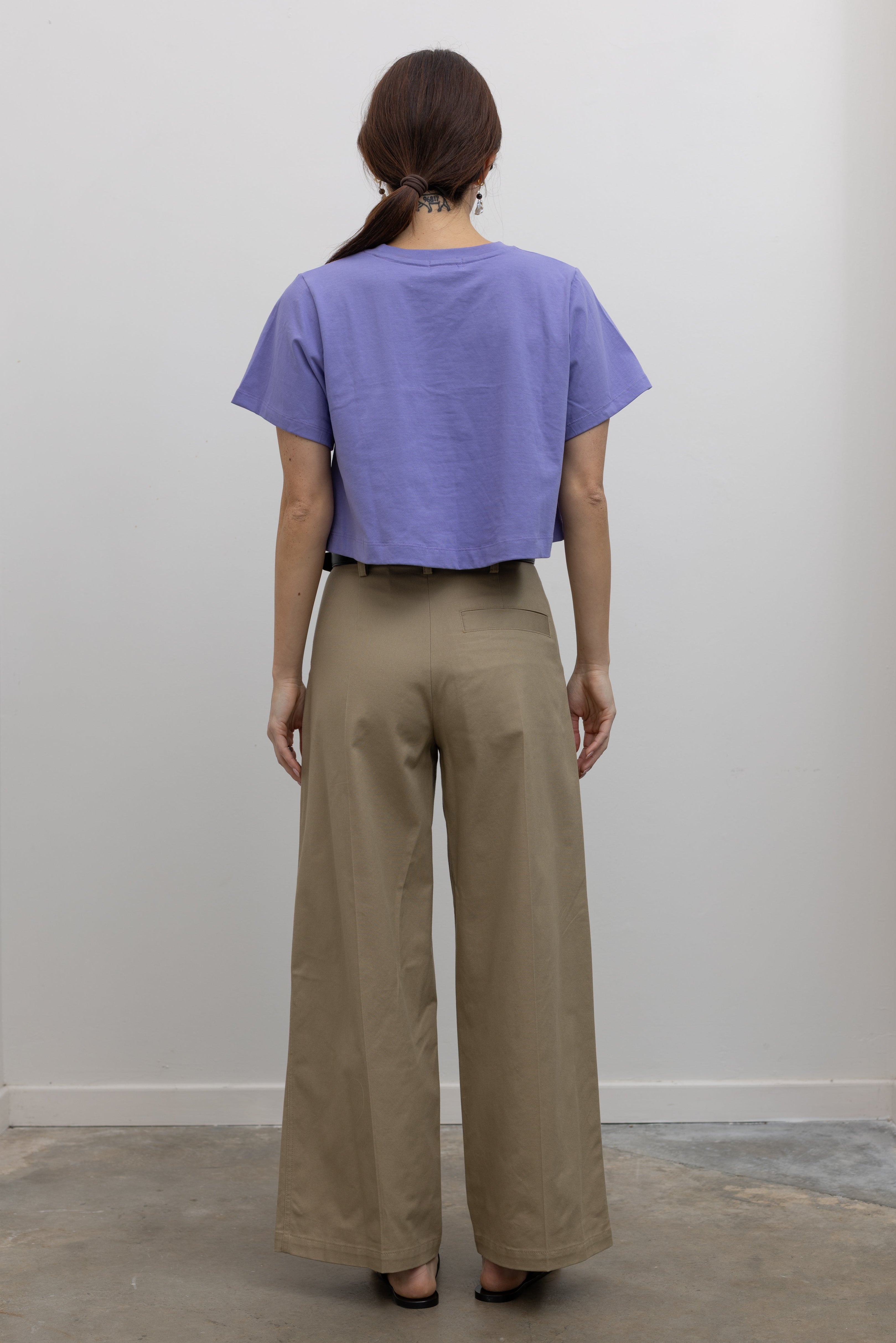 Back view of a Woman standing wearing must-have capsule wardrobe essential in an Alas Eius Label Lavender Purple Relaxed Boxy Cropped T-Shirt with khaki pleated high waisted trousers and thin black leather belt with silver hardware and black leather cutout sandals at Alas Eius 