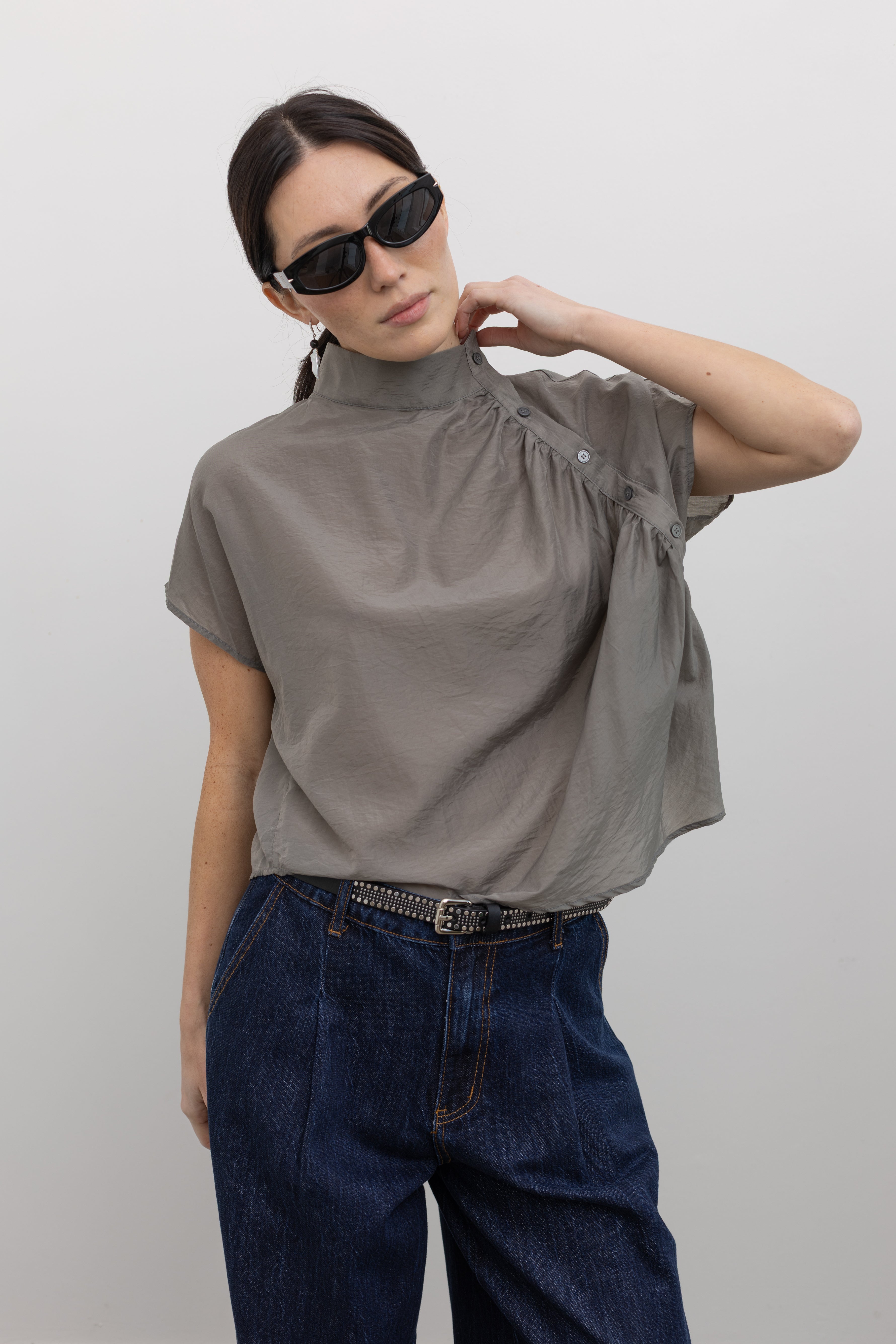 Front view of a woman wearing flattering and unique spring fashion in an Alas Eius label light 
gray organza asymmetrical button mock neck top with dark wash denim jeans at an Alas Eius store. 