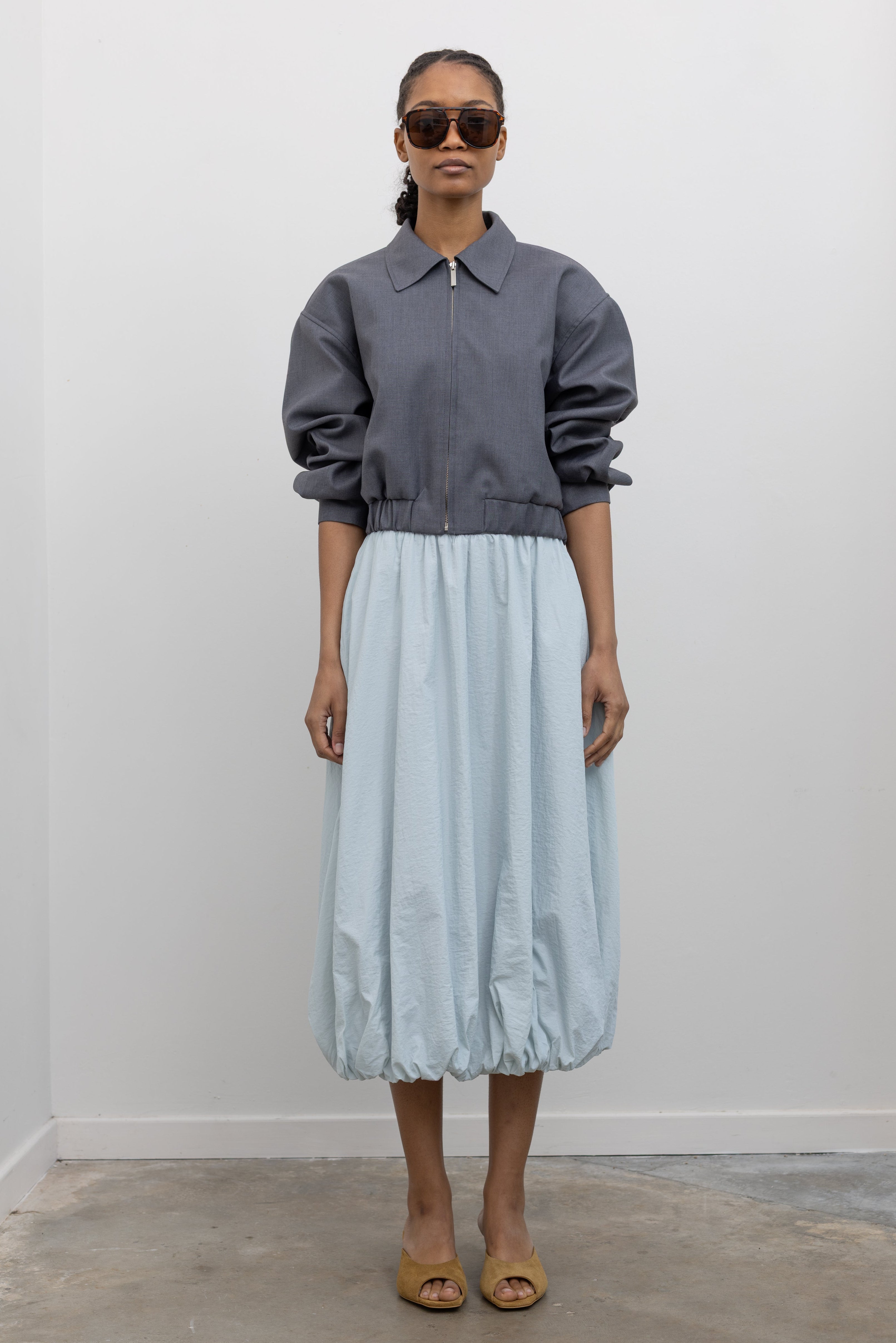Front view of a woman wearing modern minimalist womenswear spring fashion in an Alas Eius label charcoal gray cropped tailored zip jacket featuring an elastic hem for a flattering tailored look at an Alas Eius store. 