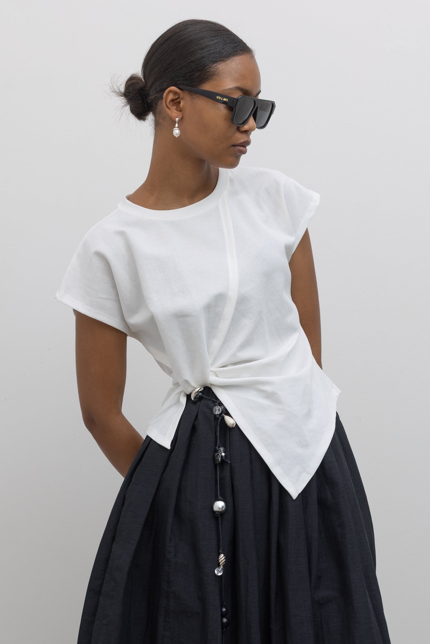 Woman wearing capsule wardrobe collection essential in an Alas Eius label White Knotted Asymmetrical Cotton Top with a black pleated skirt and a vintage beaded belt with slouchy black leather heeled boots at alas eius