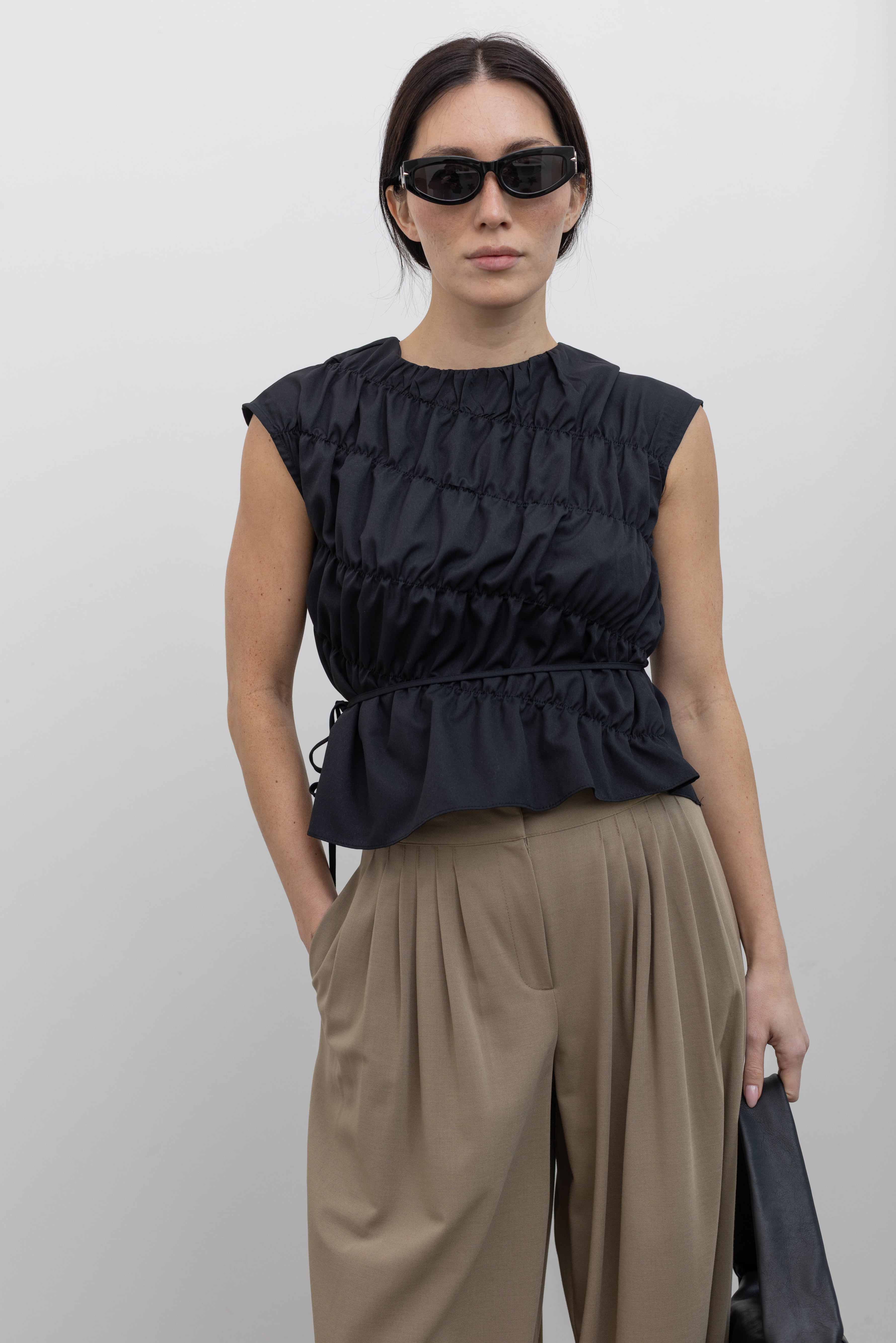 Front close-up view of a woman posing wearing contemporary and chic women's fashion in an Alas Eius Black Asymmetrical Cinched Top featuring a crew neckline, cap sleeves, and an optional waist tie paired with elevated khaki cinched ankle pants, silver sandals, and a black leather St. Agni bag at an Alas Eius store. 