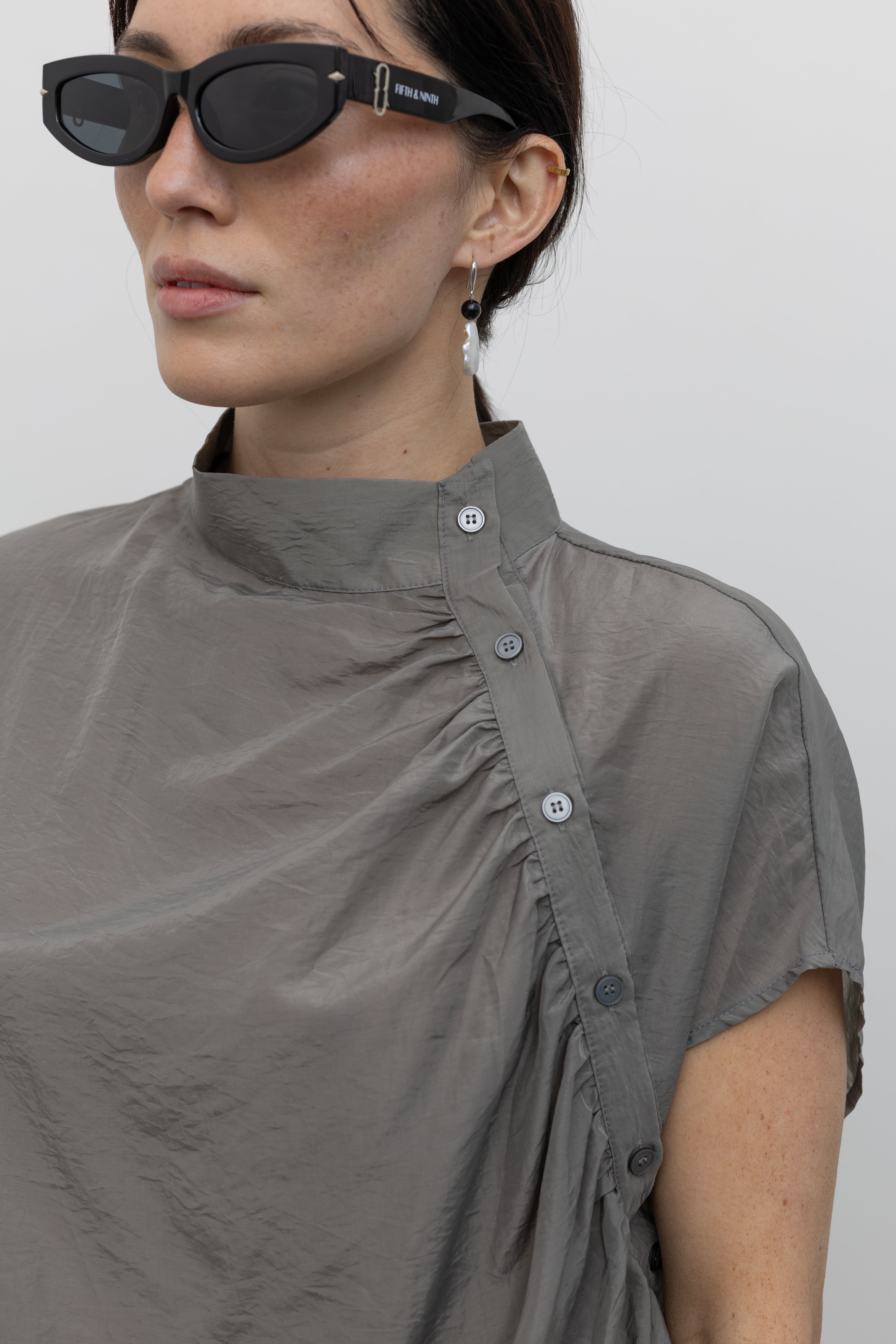 Close-up front view of a woman wearing modern minimalist spring fashion in an Alas Eius label light 
gray organza diagonal button front mock neck top with dark wash denim jeans at an Alas Eius store. 