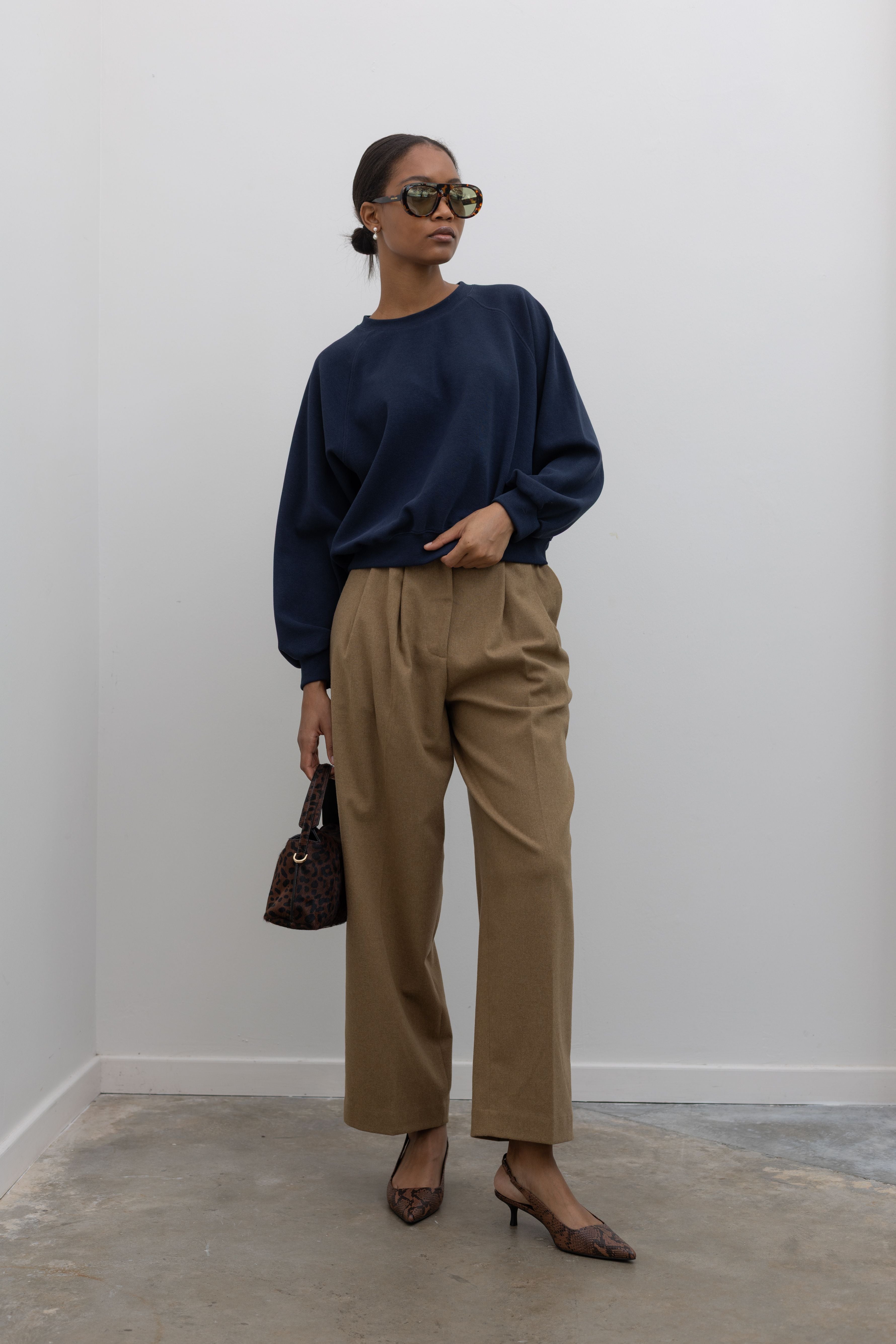 Full front view of a woman standing wearing a capsule wardrobe essential style in an Alas Eius label navy blue raglan sleeve elevated sweatshirt with a slightly cropped hem, landing at the waistband. Paired with tan trousers, animal print slingback kitten heels and sunglasses at Alas Eius. 