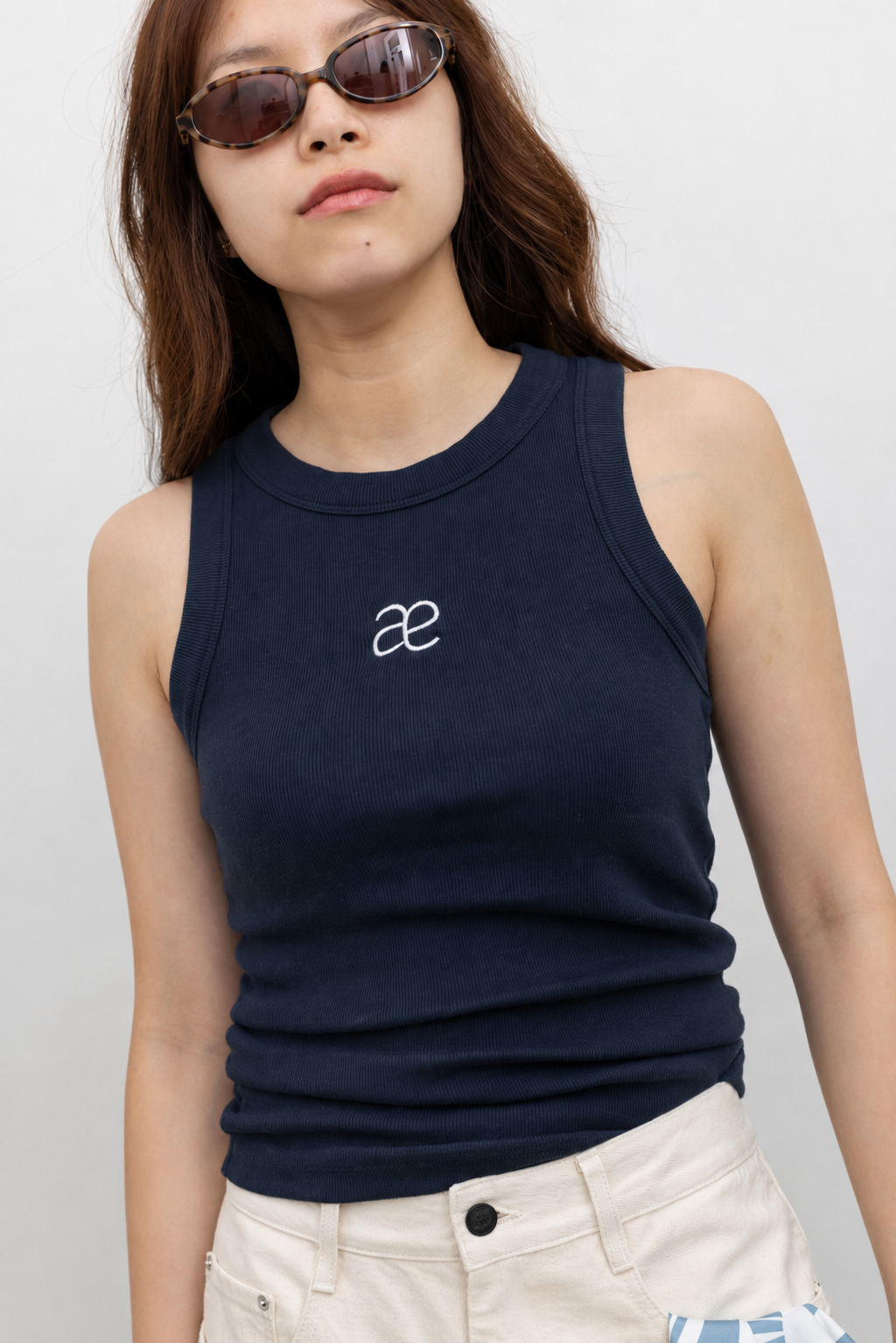 Model wearing effortless style in a Alas Eius label navy blue cotton blend ribbed racerback tank top with contrasting embroidered AE logo on chest paired Collective Ivory Balloon Curved Cotton Pant at Alas Eius.