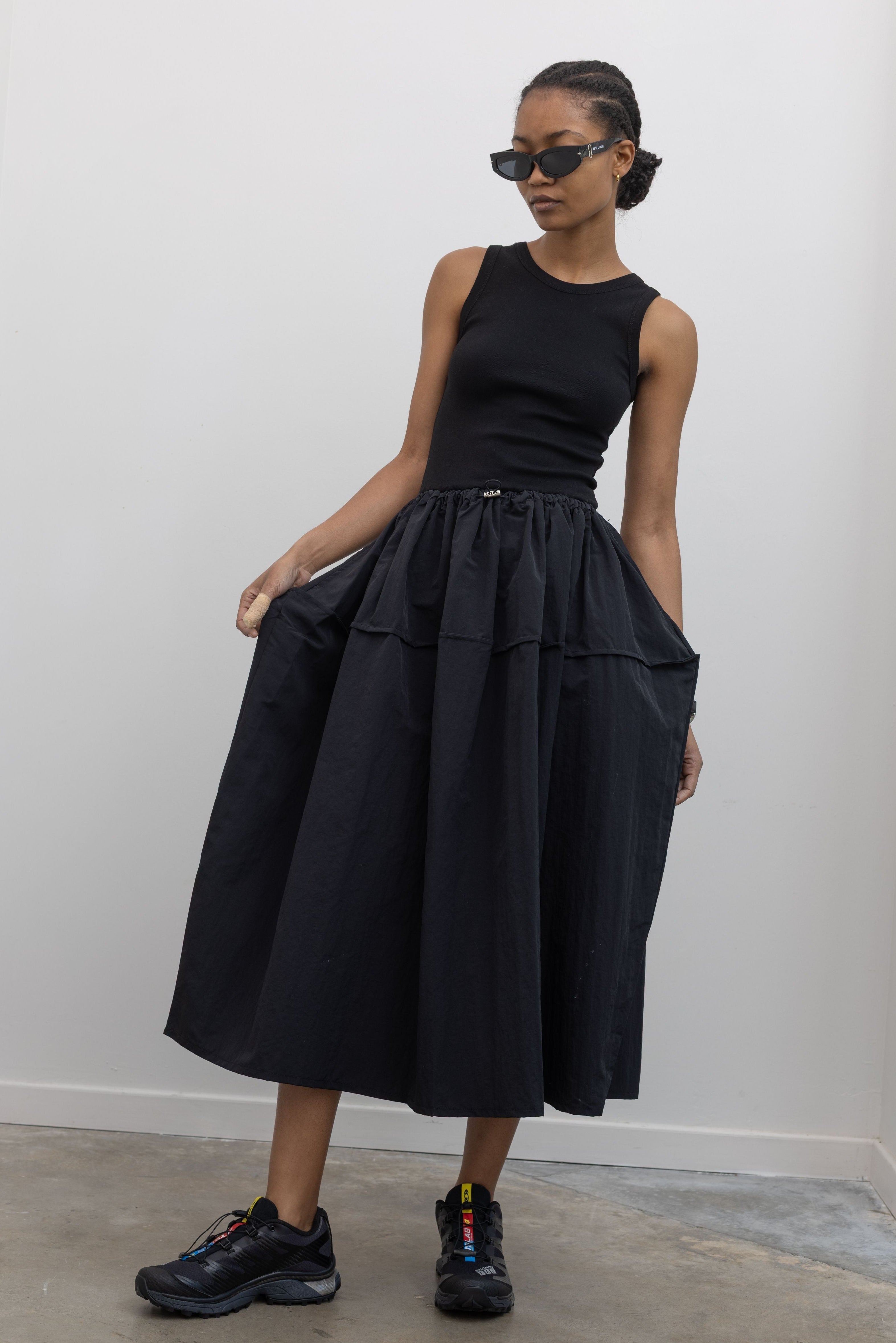 Woman wearing Spring fashion in a flattering Alas Eius brand dress with a  cotton bodice and a structured nylon skirt cinched by an adjustable drawstring waist paired with black Salomon sneakers and sunglasses at Alas Eius 