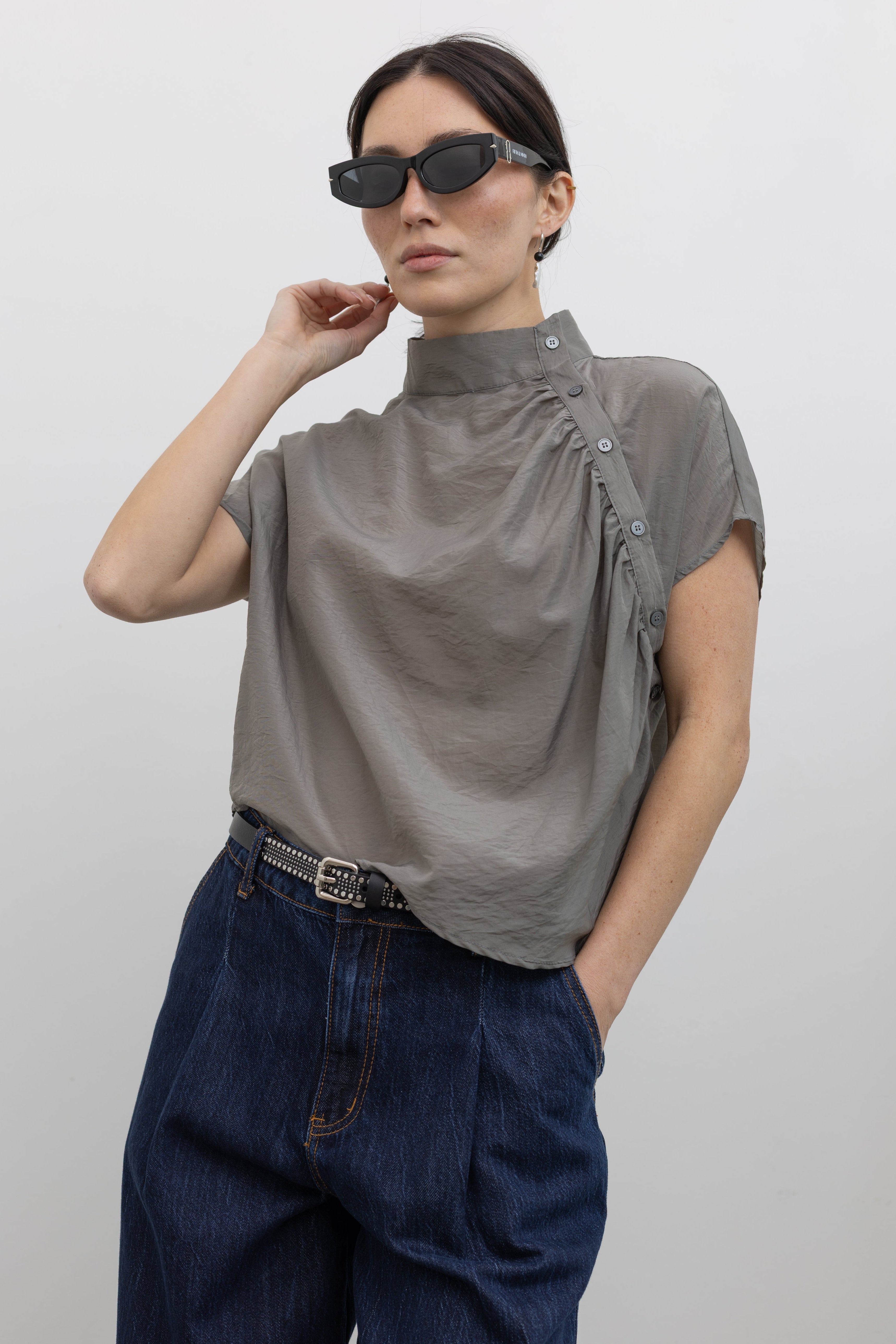 Woman wearing contemporary spring fashion in an Alas Eius label light 
gray organza asymmetrical button mock neck top with dark wash denim jeans at an Alas Eius store. 