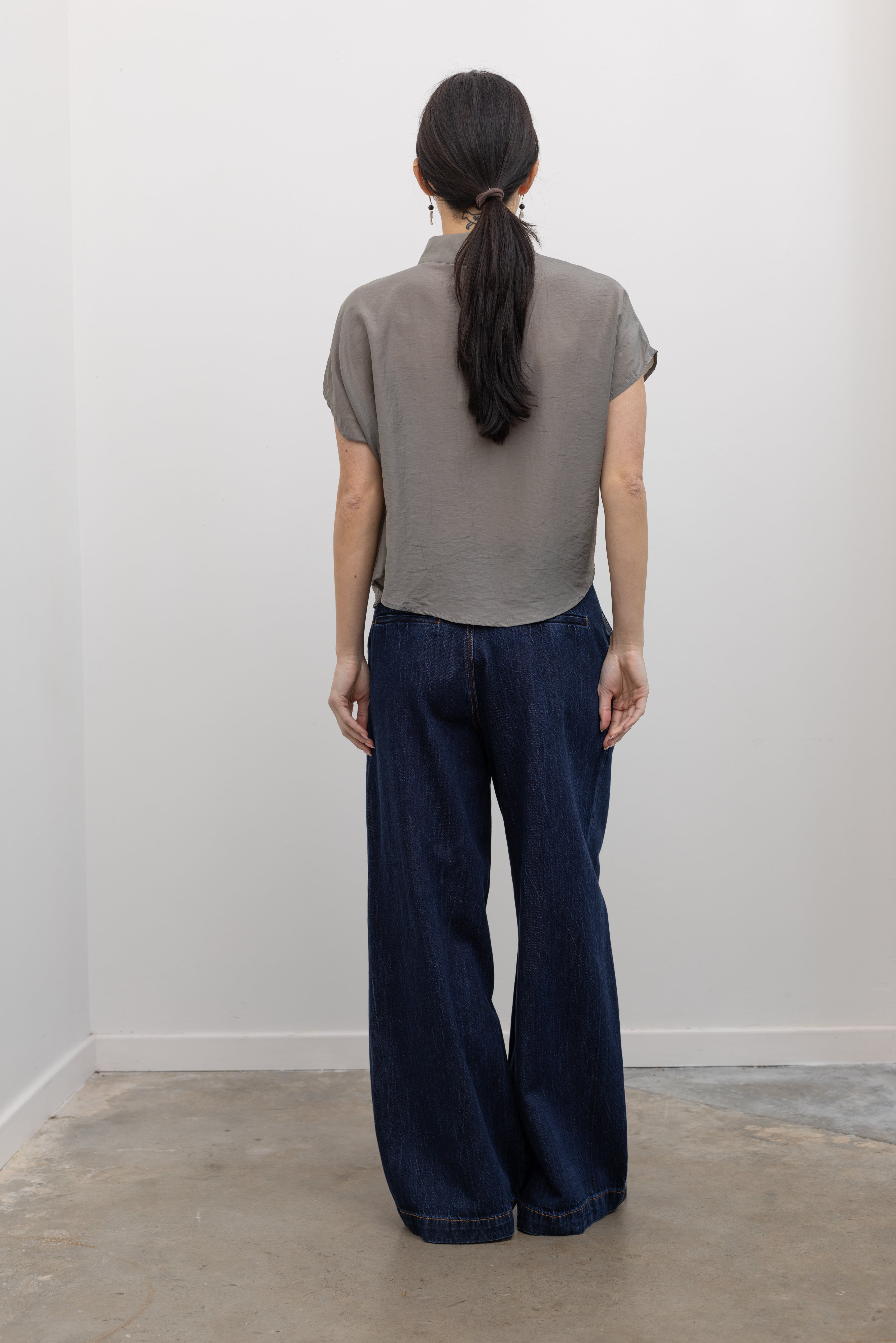 Full length back view of a woman wearing contemporary spring fashion in an Alas Eius label light 
gray organza asymmetrical button mock neck top with dark wash denim jeans at an Alas Eius store. 