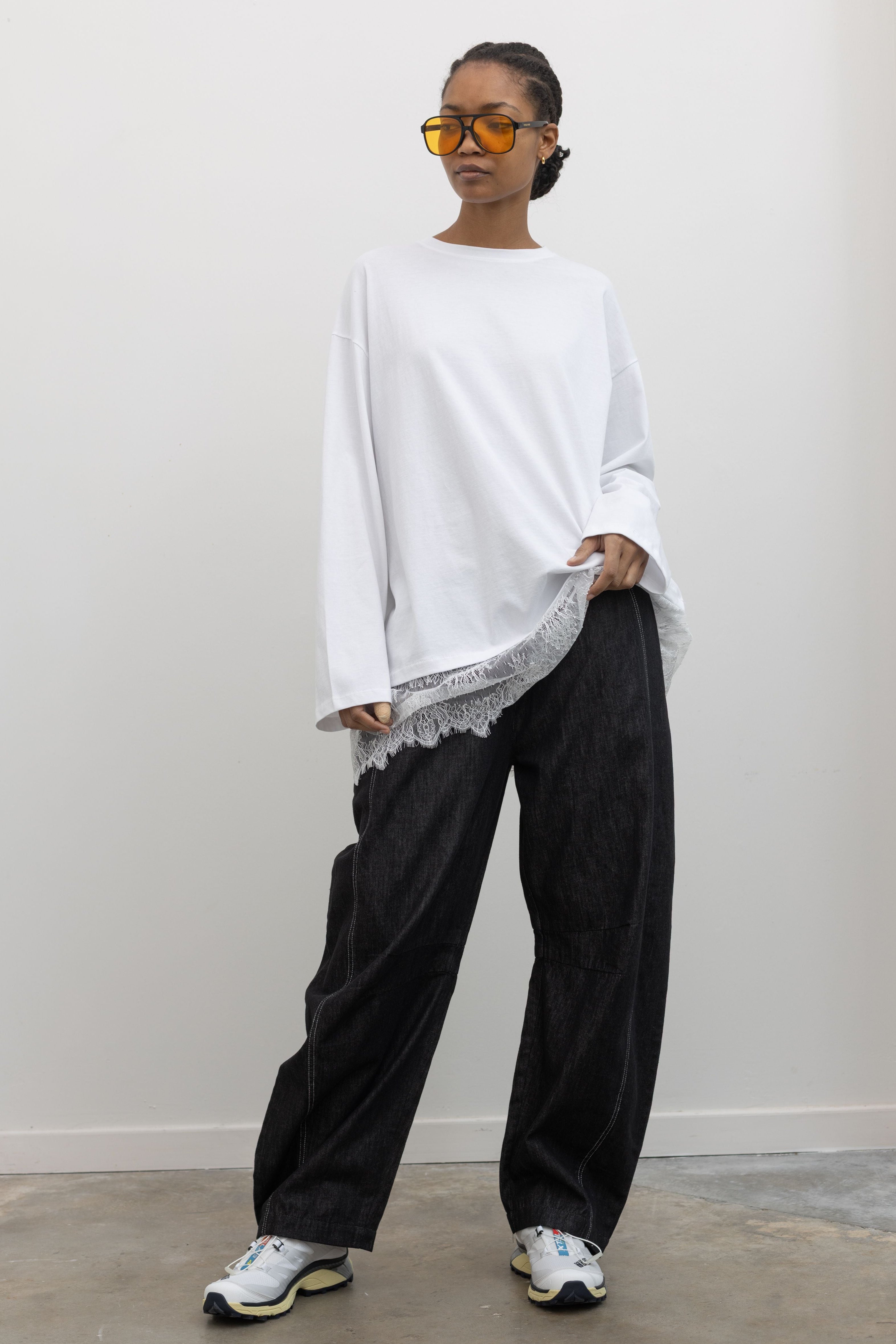 Full length front view of a woman posing wearing elevated spring fashion in a feminine Alas Eius label white long-sleeve oversized tunic top with lace hemline at and Alas Eius store. 