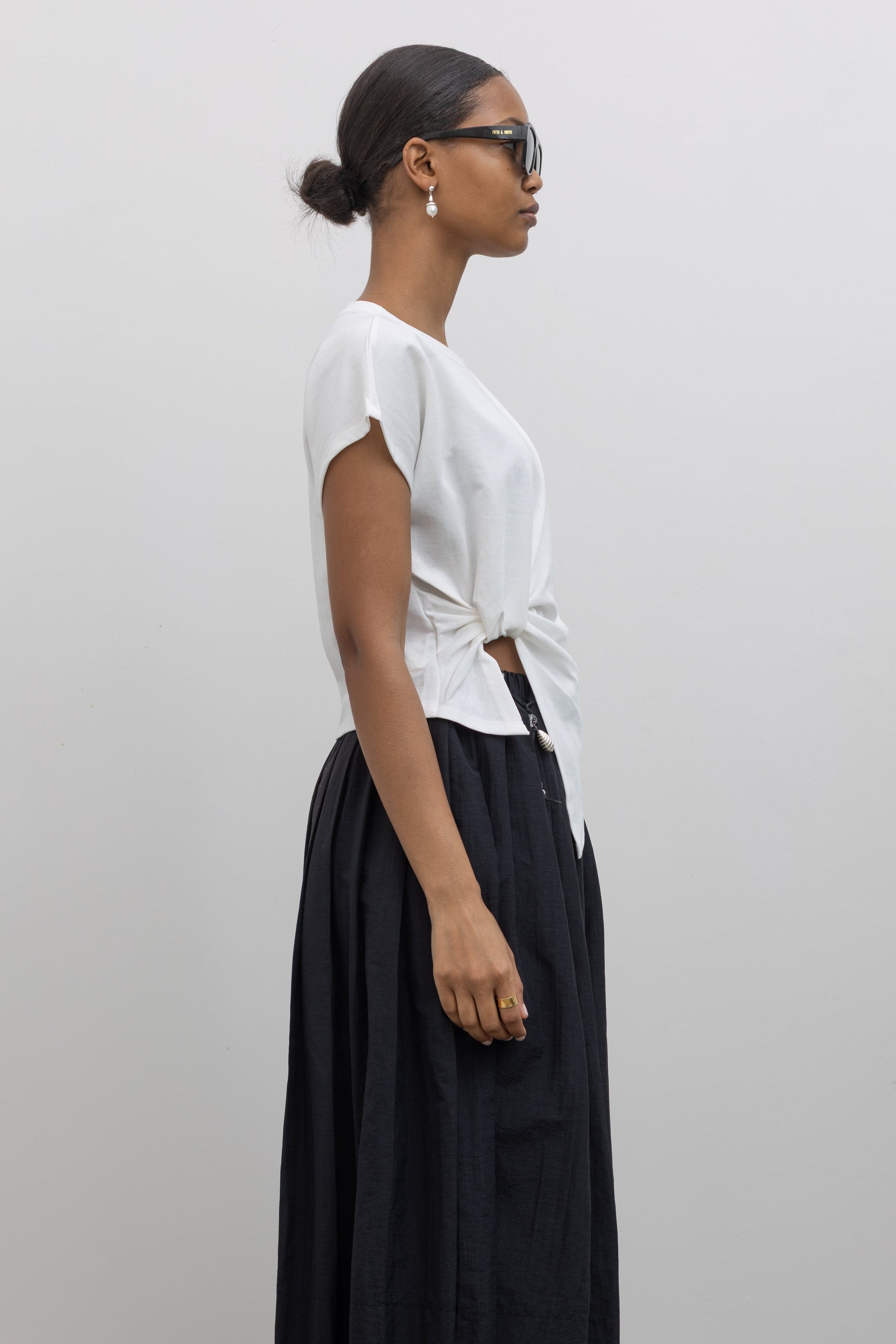 Side view Woman wearing everyday capsule wardrobe collection essential in an Alas Eius label White Knotted Asymmetrical Cotton Top with a black pleated skirt and a vintage beaded belt at alas eius