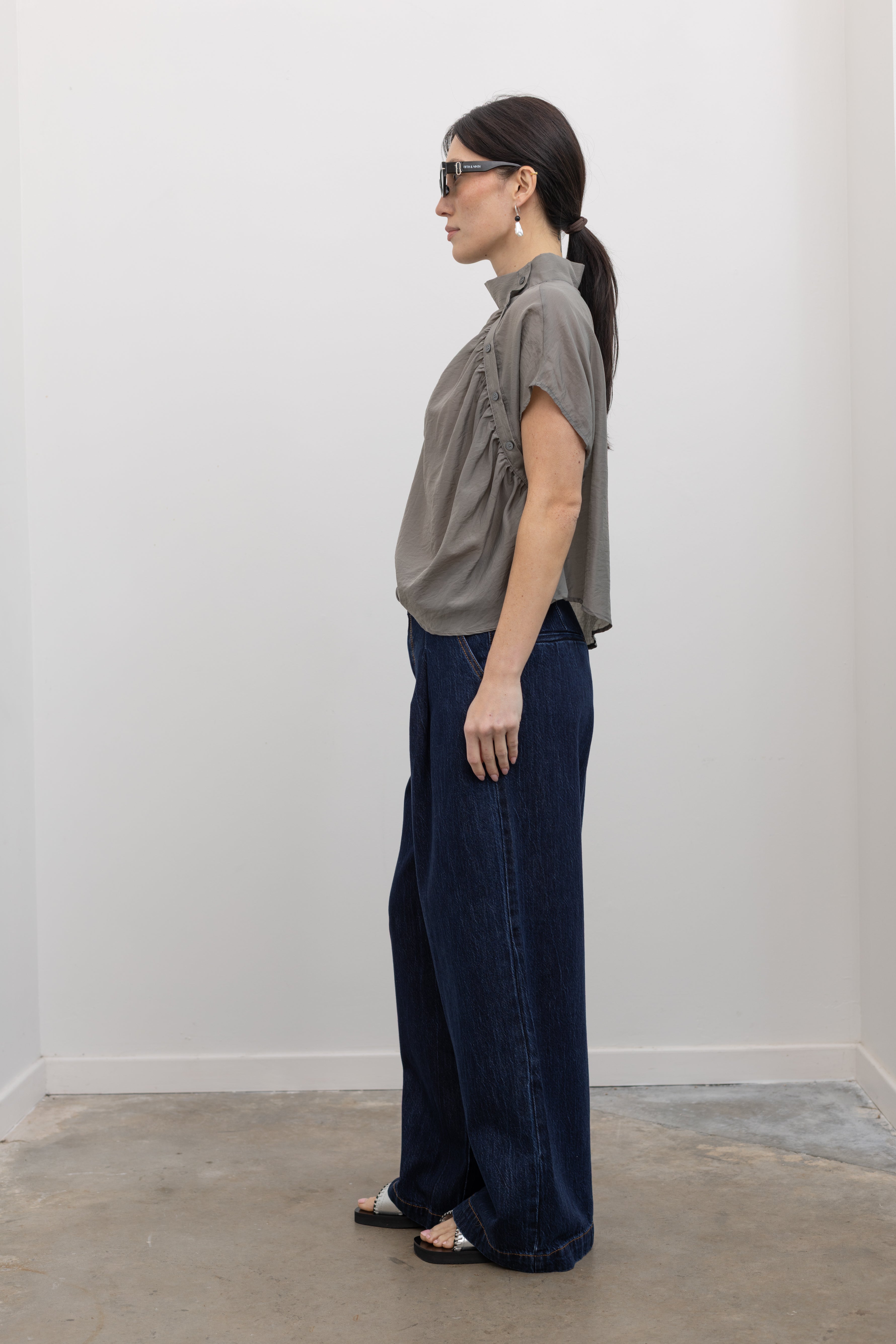 Full length side view of a woman wearing contemporary spring fashion in an Alas Eius label light 
gray organza asymmetrical button mock neck top with dark wash denim jeans at an Alas Eius store. 