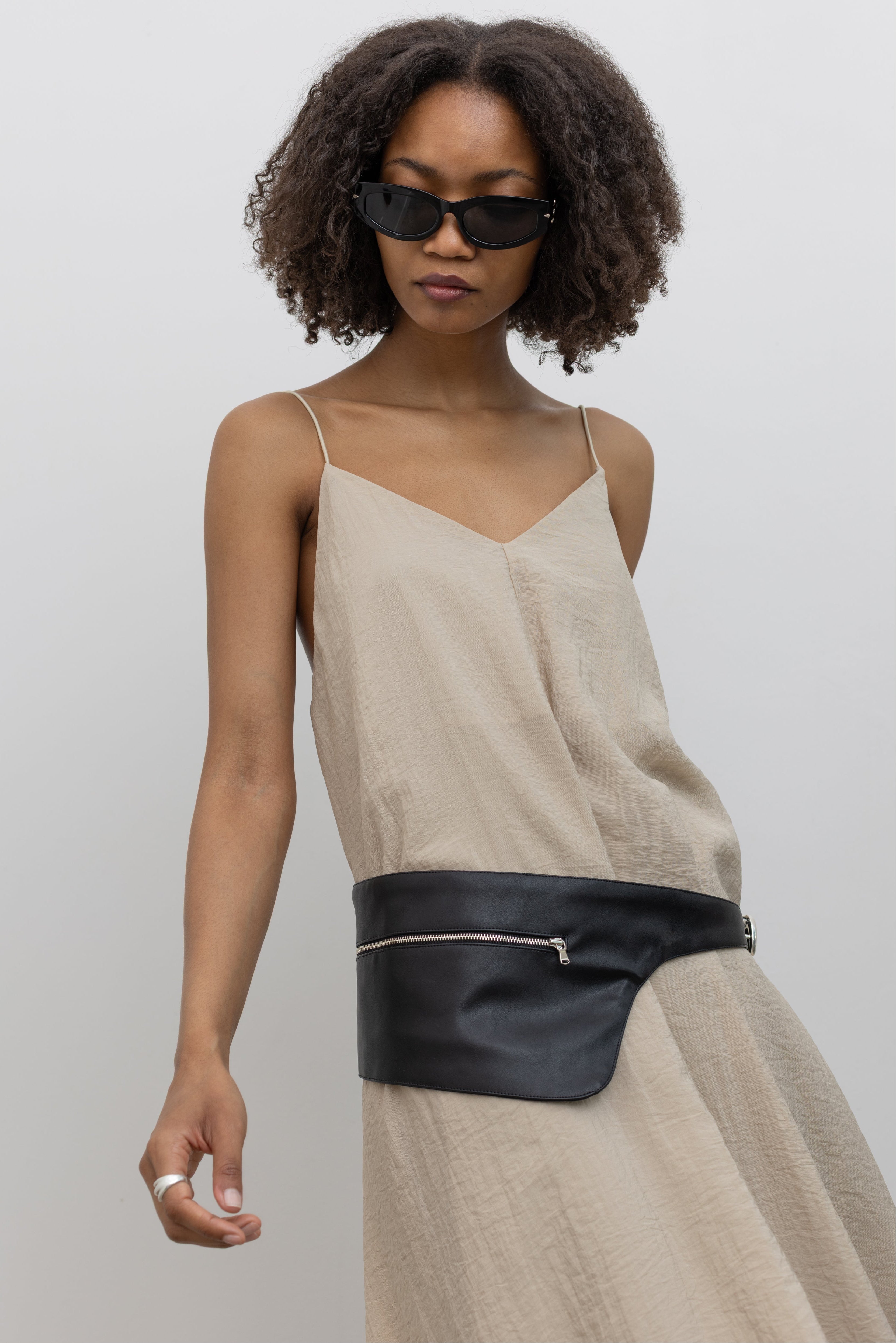 Front view of a woman wearing an elegant yet minimalist Alas Eius label spring and summer dress in a beige cotton blend featuring a flowing silhouette, adjustable bungee straps for a customized fit and two convenient side pockets paired with Nomia black leather zip belt bag around her waist at an Alas Eius store. 