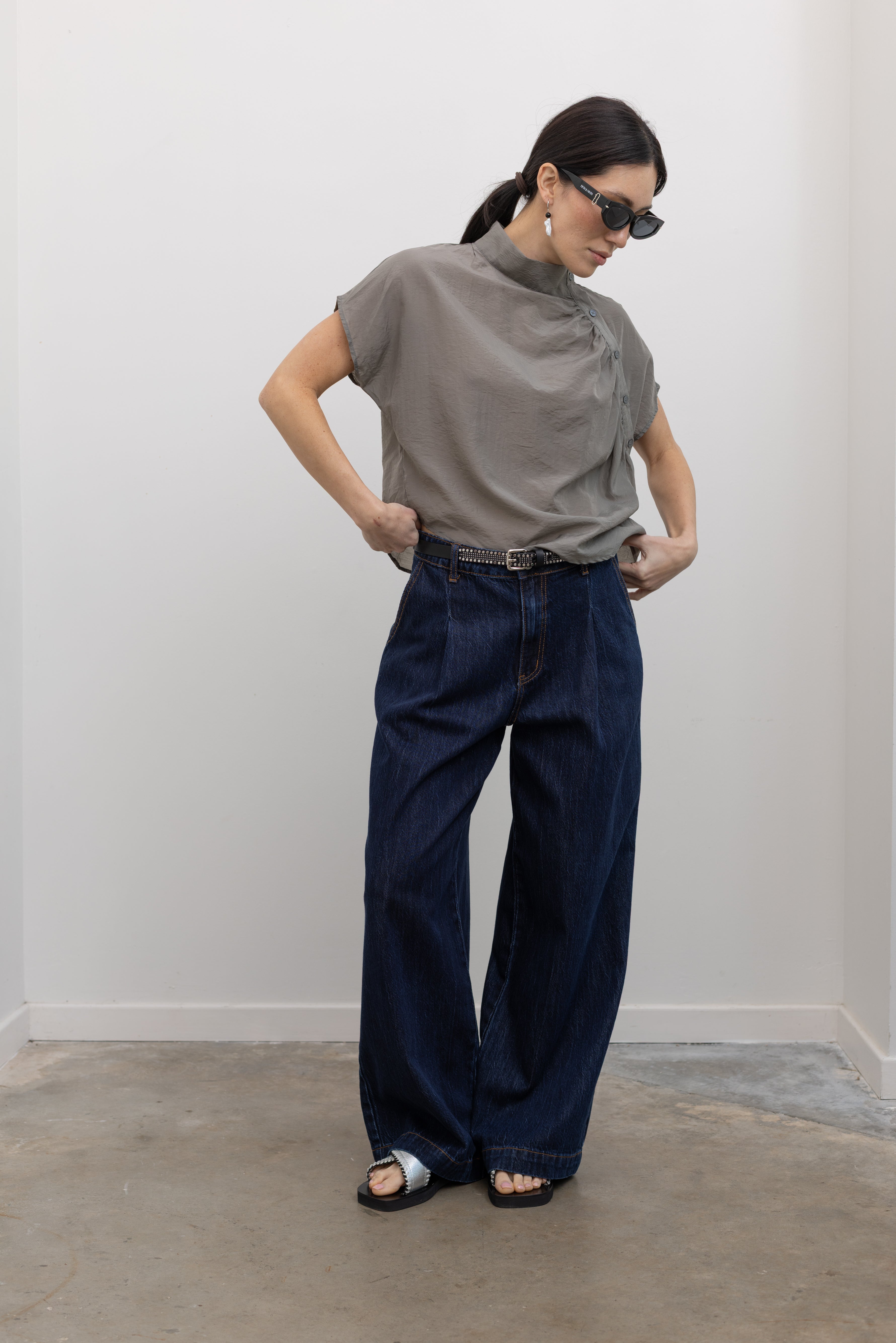 Full length front view of a woman wearing contemporary spring fashion in an Alas Eius label light 
gray organza asymmetrical button mock neck top with dark wash denim jeans at an Alas Eius store. 