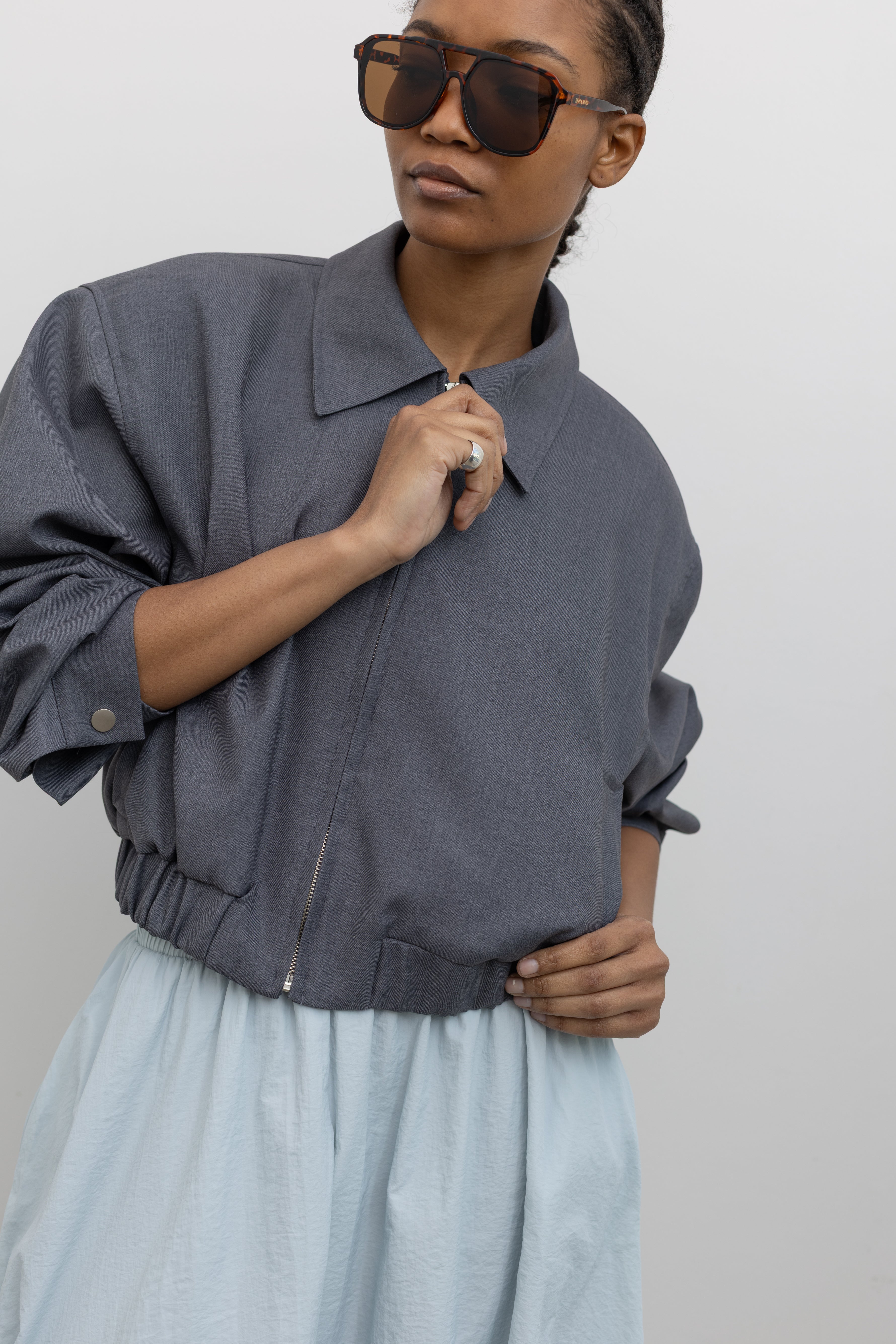Front view of a woman wearing versatile and refined womenswear spring fashion in an Alas Eius label charcoal gray cropped tailored zip jacket featuring an elastic hem for a flattering cinched waist at an Alas Eius store. 