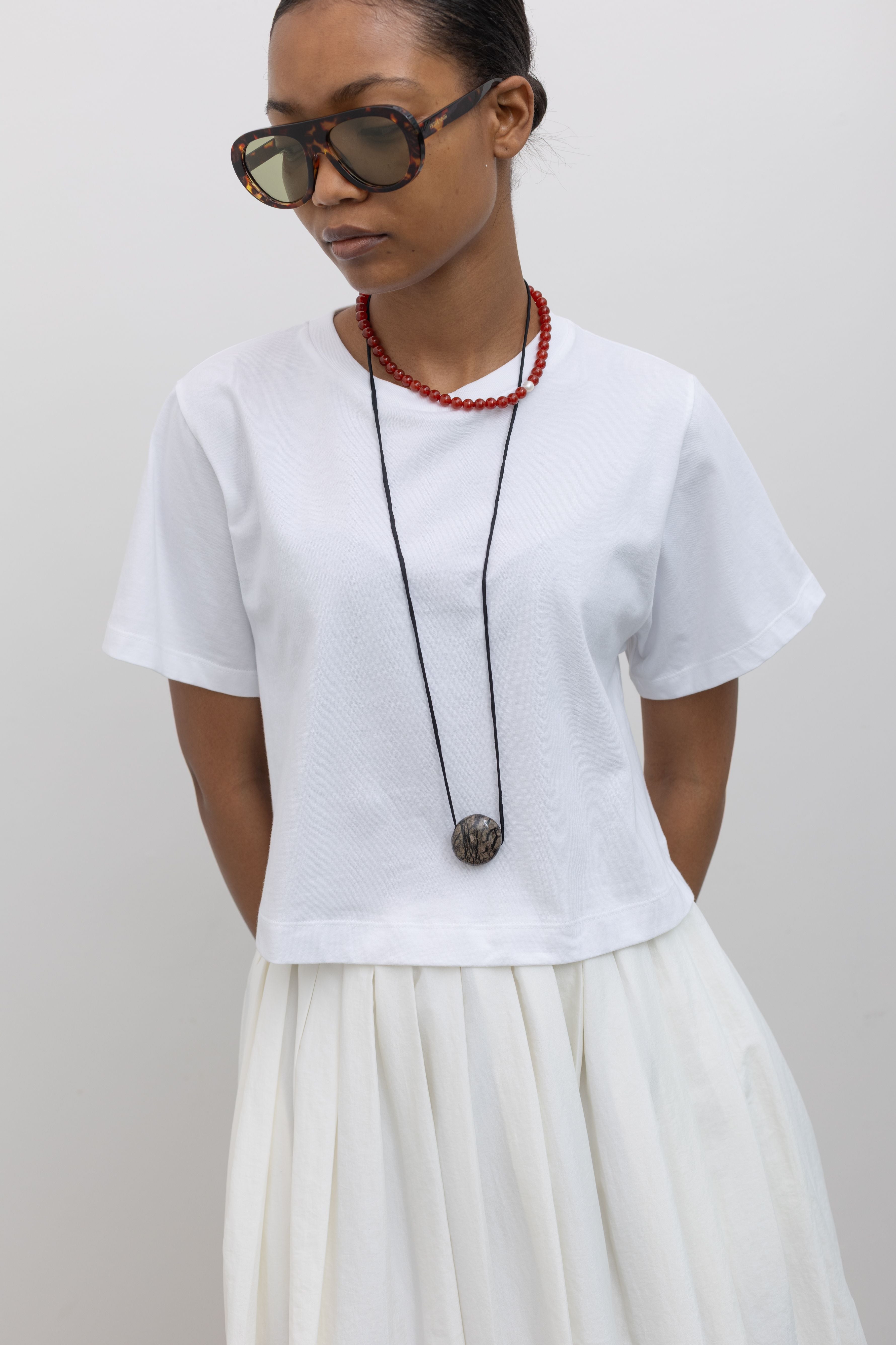 Detailed product view of woman standing wearing everyday essential Alas Eius Label white cotton Relaxed Cropped Short Sleeved T-Shirt with sunglasses and white nylon pleated midi skirt at Alas Eius