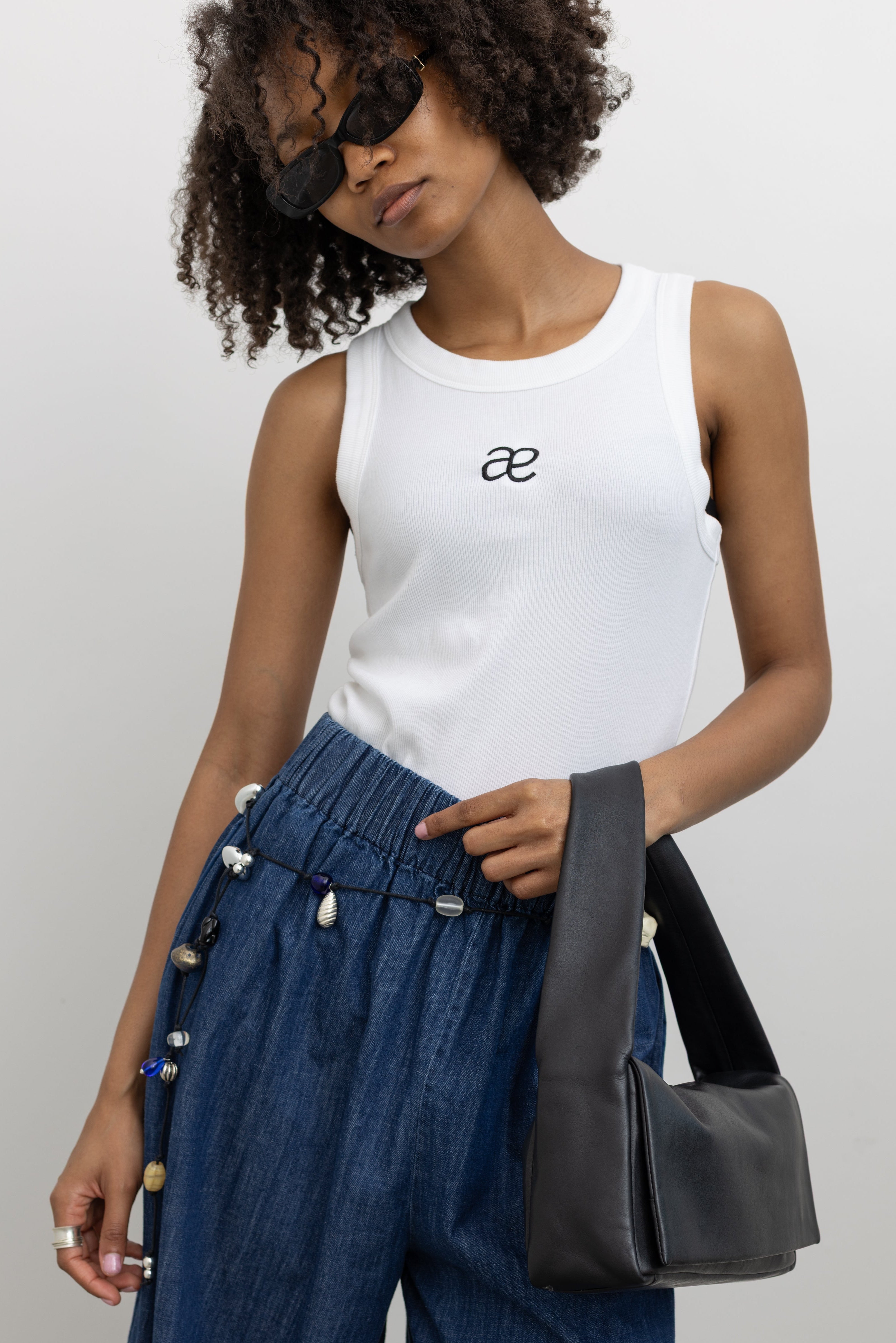 Model wearing an everyday essential style in an Alas Eius label white cotton blend ribbed racerback tank top with contrasting embroidered AE logo on chest paired Collective blue cotton denim wide leg flowy pull on pant at Alas Eius. 