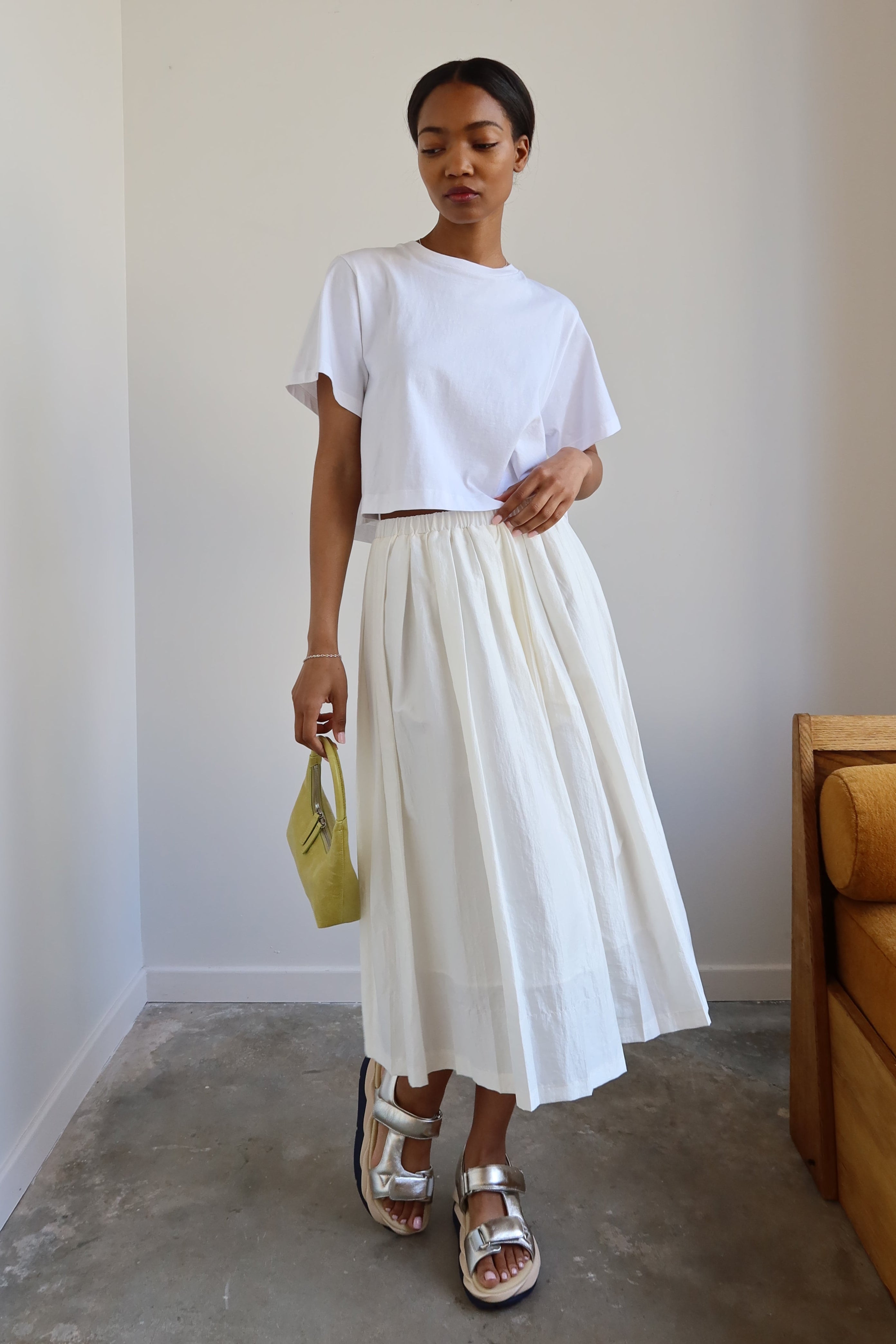 A woman wearing a white t-shirt and a white pleated full skirt with an elastic waistband and side pockets. She is also wearing sandals and carrying a small yellow purse.
