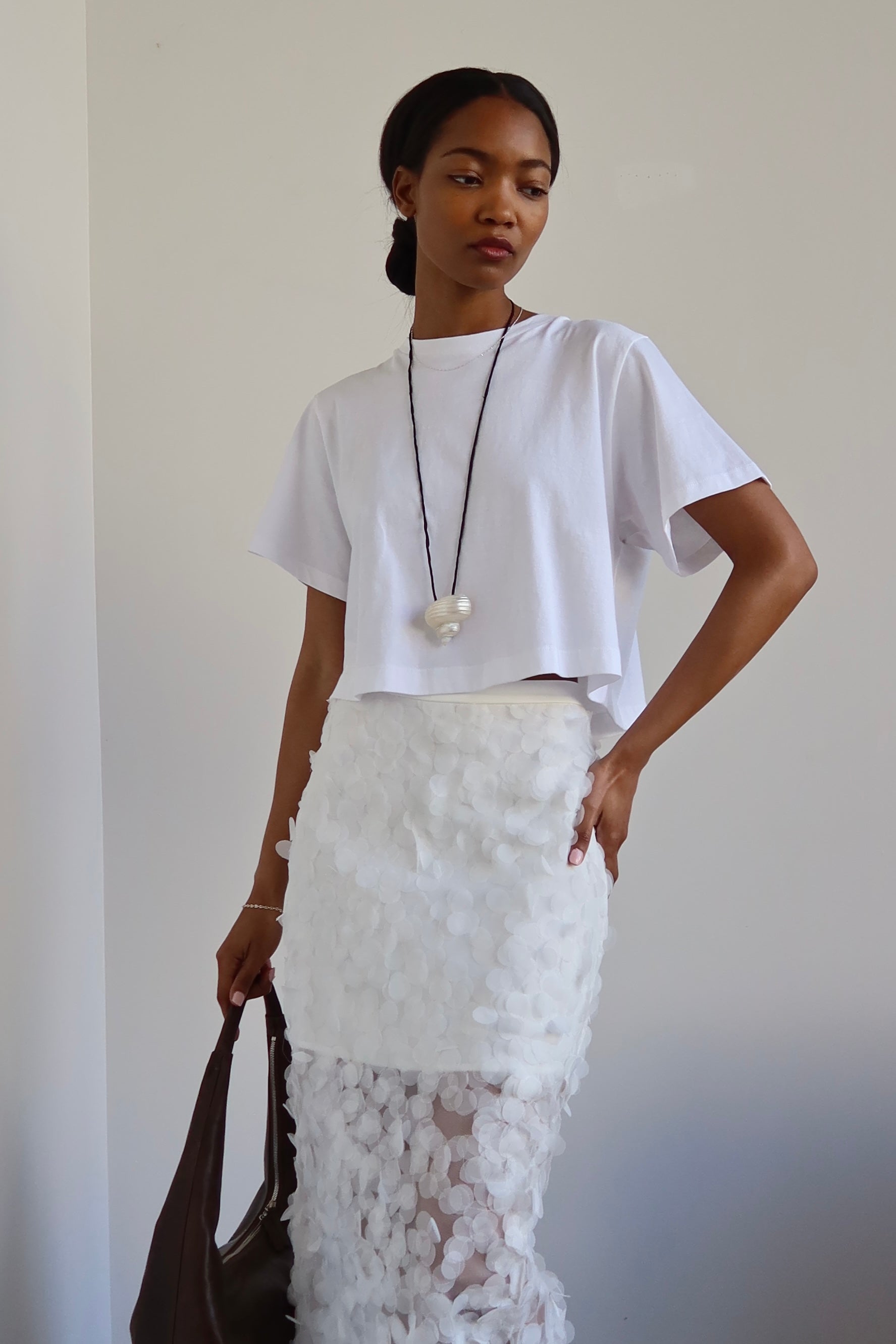 A woman wearing a white relaxed fit cropped t-shirt paired with a long white skirt, accessorized with a necklace and carrying a brown bag.