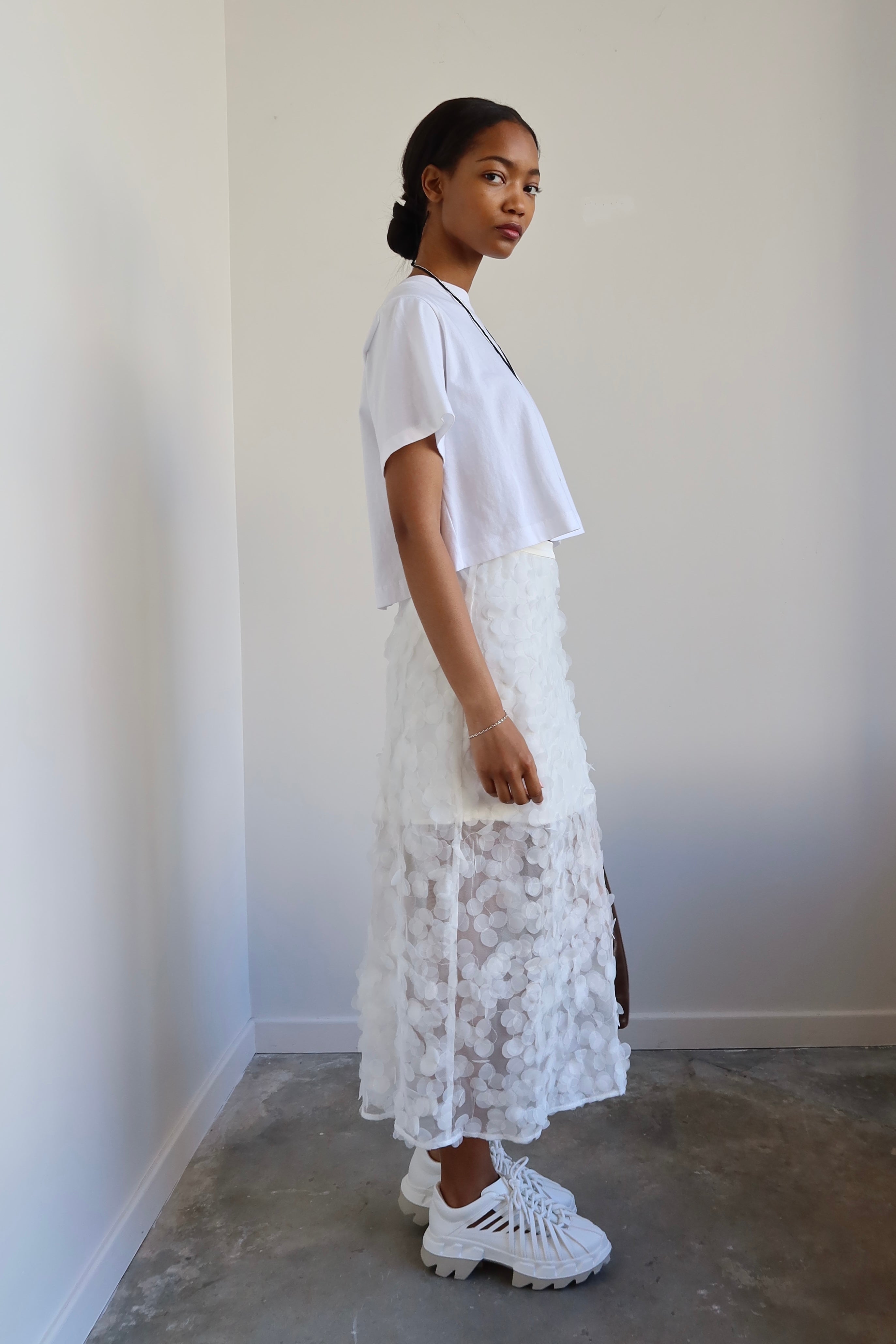 A woman wearing a white relaxed fit cropped t-shirt paired with a long white skirt, accessorized with a necklace and carrying a brown bag.