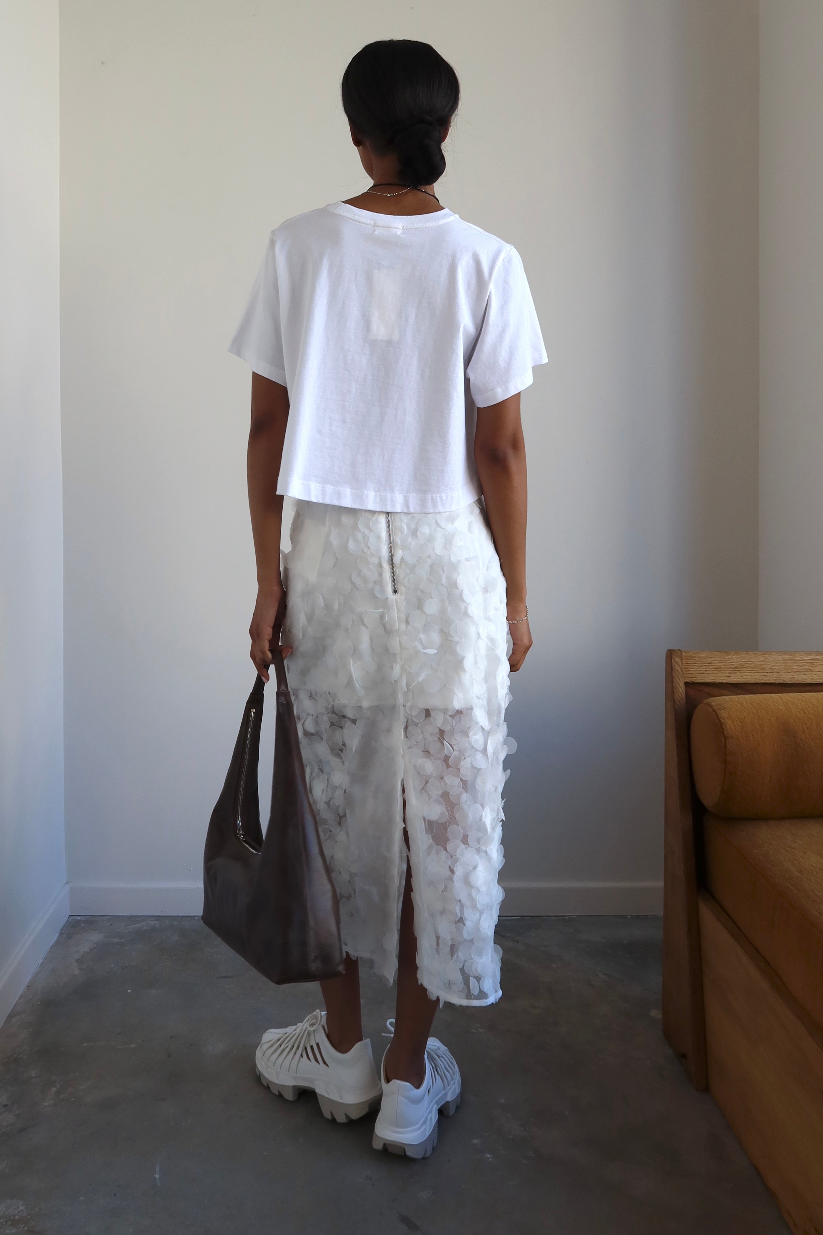 A woman wearing a white relaxed fit cropped t-shirt paired with a long white skirt, accessorized with a necklace and carrying a brown bag.