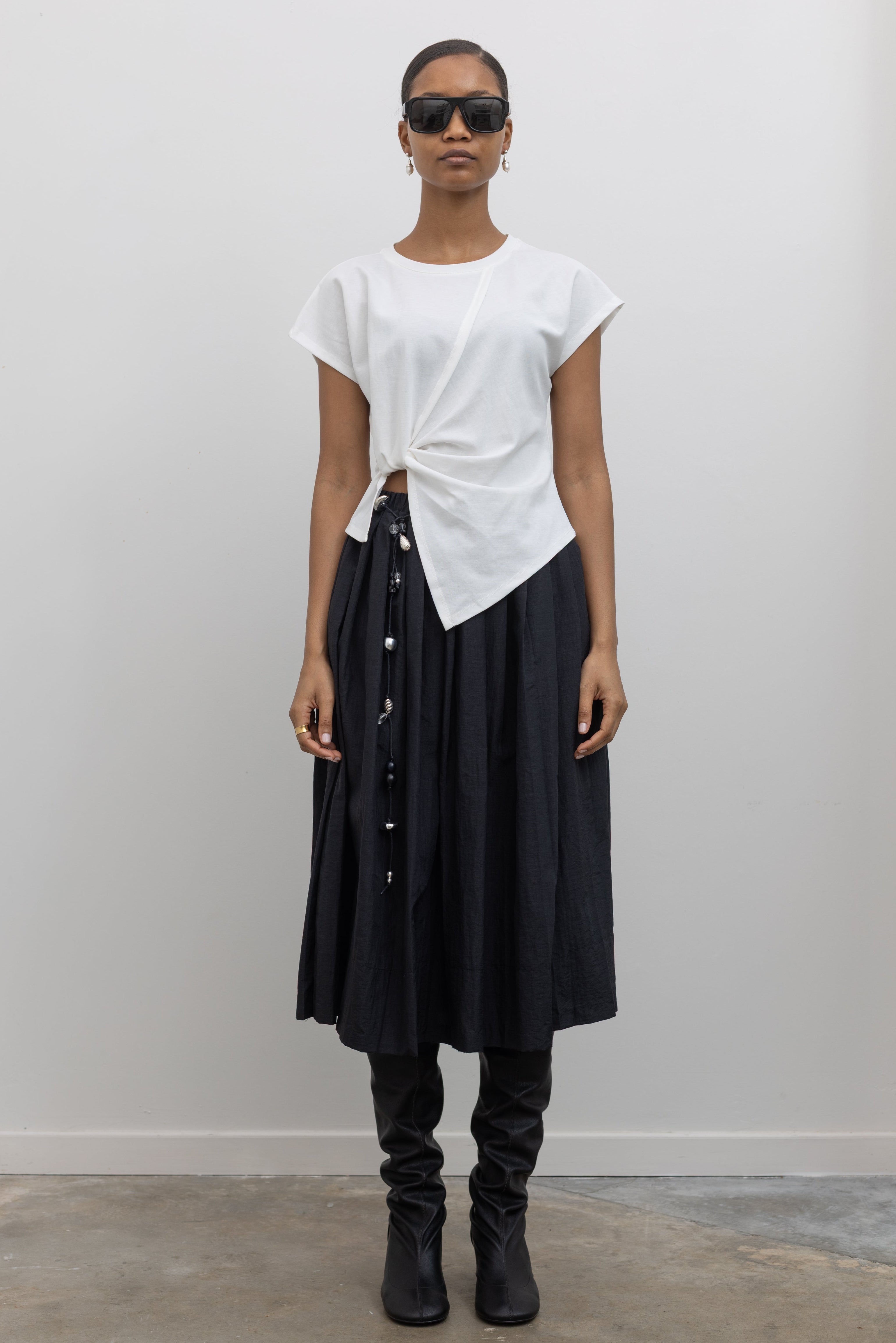 Woman wearing everyday capsule wardrobe collection essential in an Alas Eius label White Knotted Asymmetrical Cotton Top with a black pleated skirt and a vintage beaded belt and slouchy black leather heeled boots at alas eius