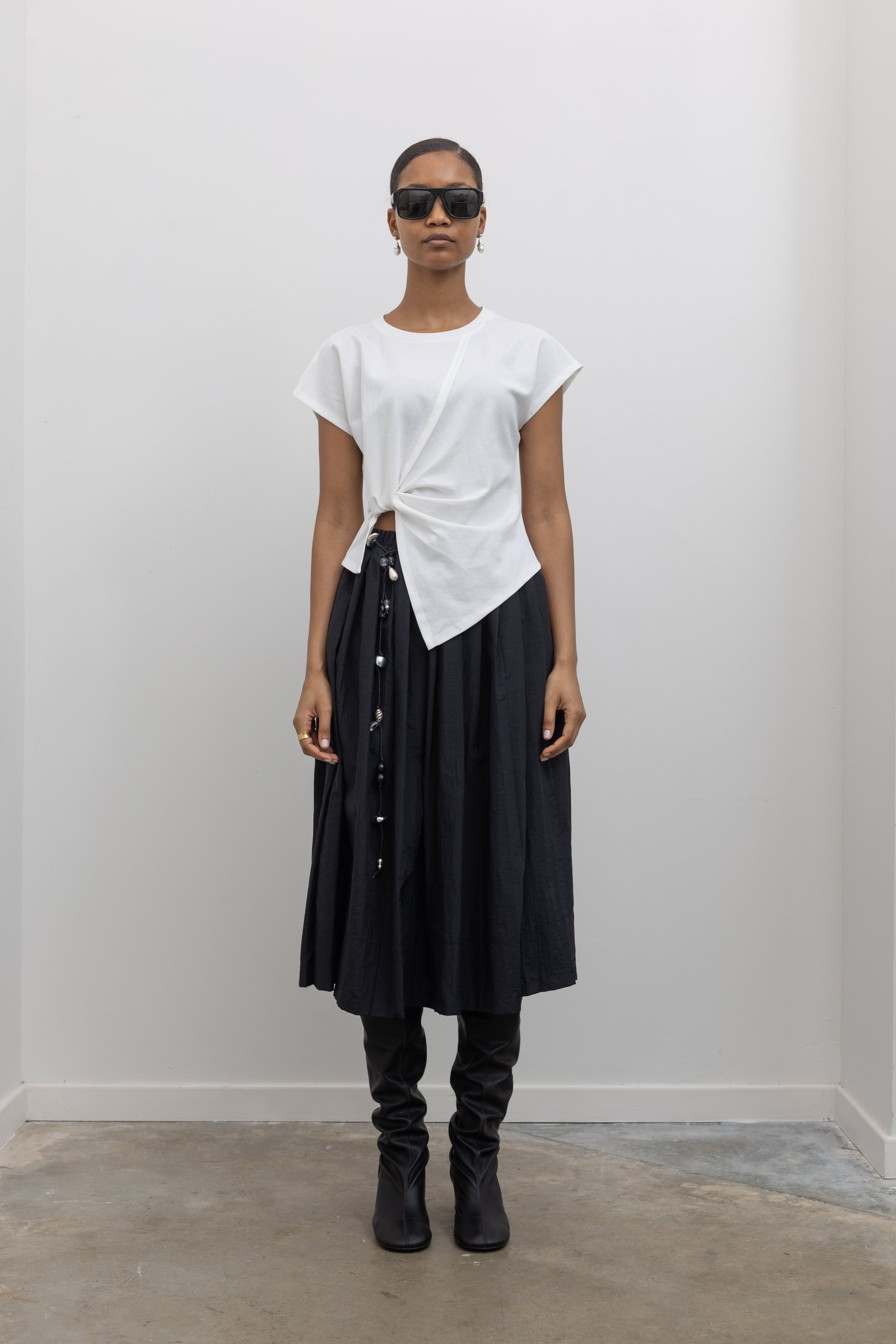 Woman wearing everyday capsule wardrobe collection essential in an Alas Eius label White Knotted Asymmetrical Cotton Top with a black pleated skirt and a vintage beaded belt and slouchy black leather heeled boots at alas eius