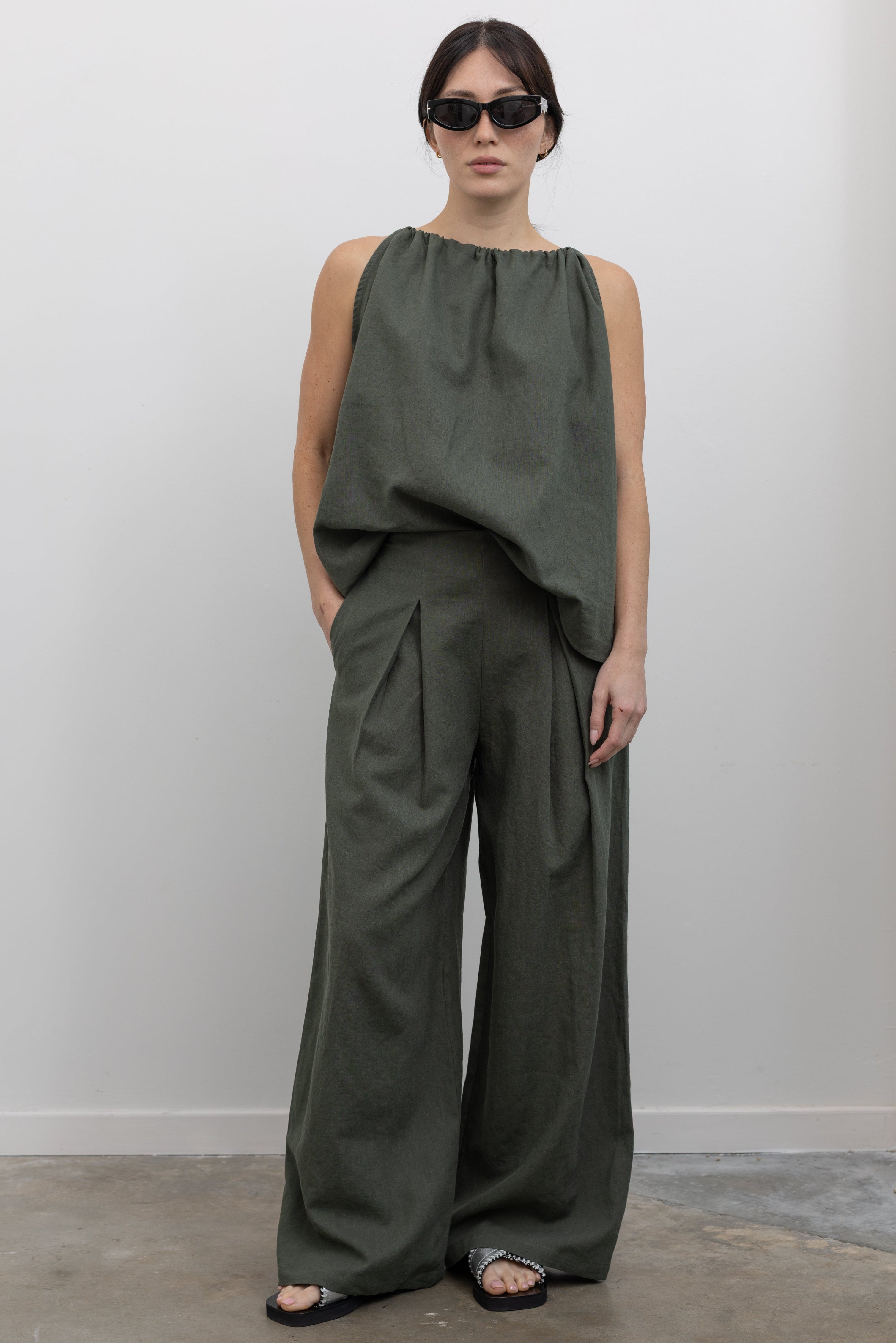 Full length front view of a woman standing wearing modern minimalist spring and summer fashion in an Alas Eius label cotton and linen blend wide leg pleated front trouser paired with its coordinating Halter Neck Cotton Top at an Alas Eius store. 