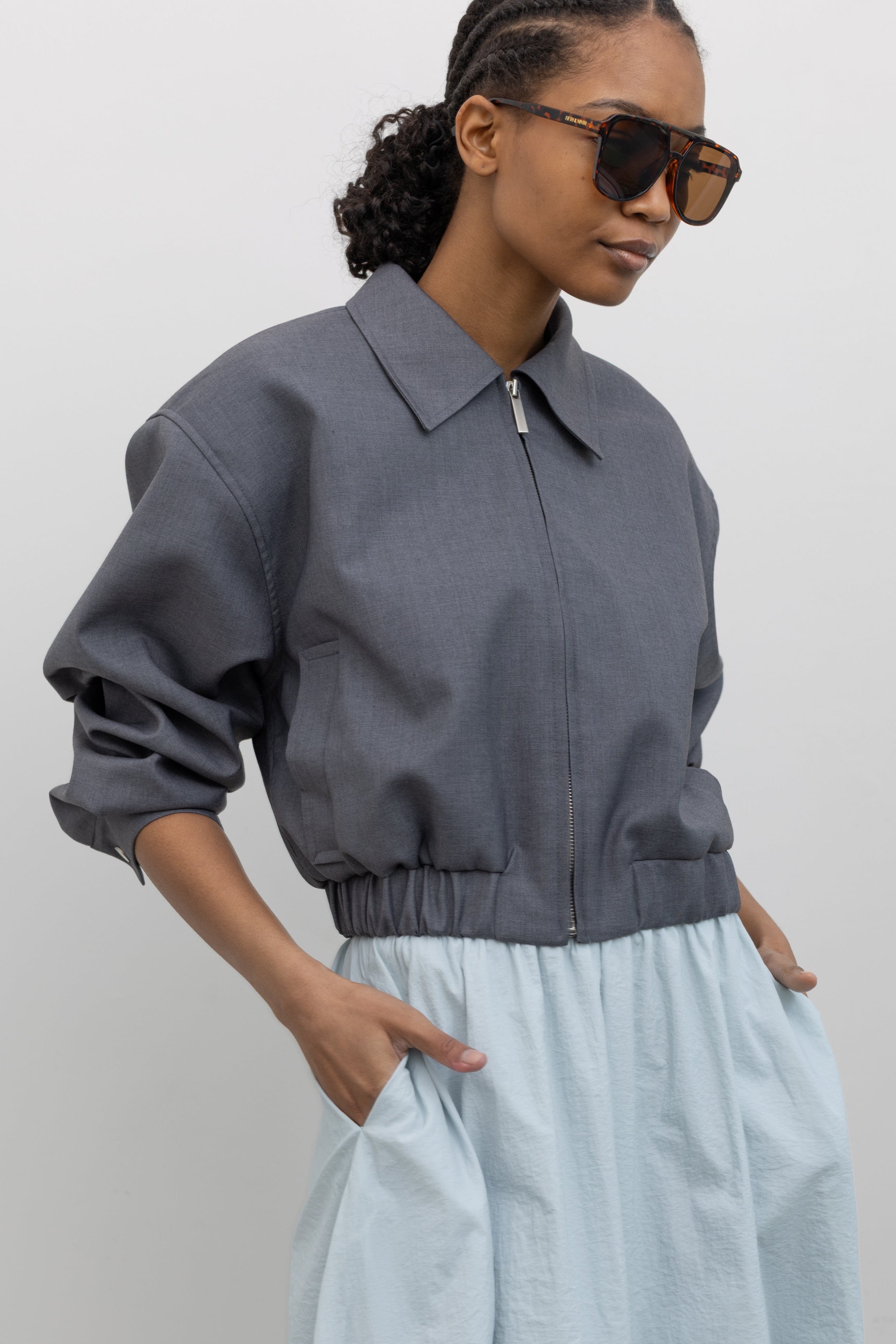 3/4 view of a woman wearing elevated spring fashion in a Alas Eius label charcoal cropped tailored zip jacket at an Alas Eius store. 