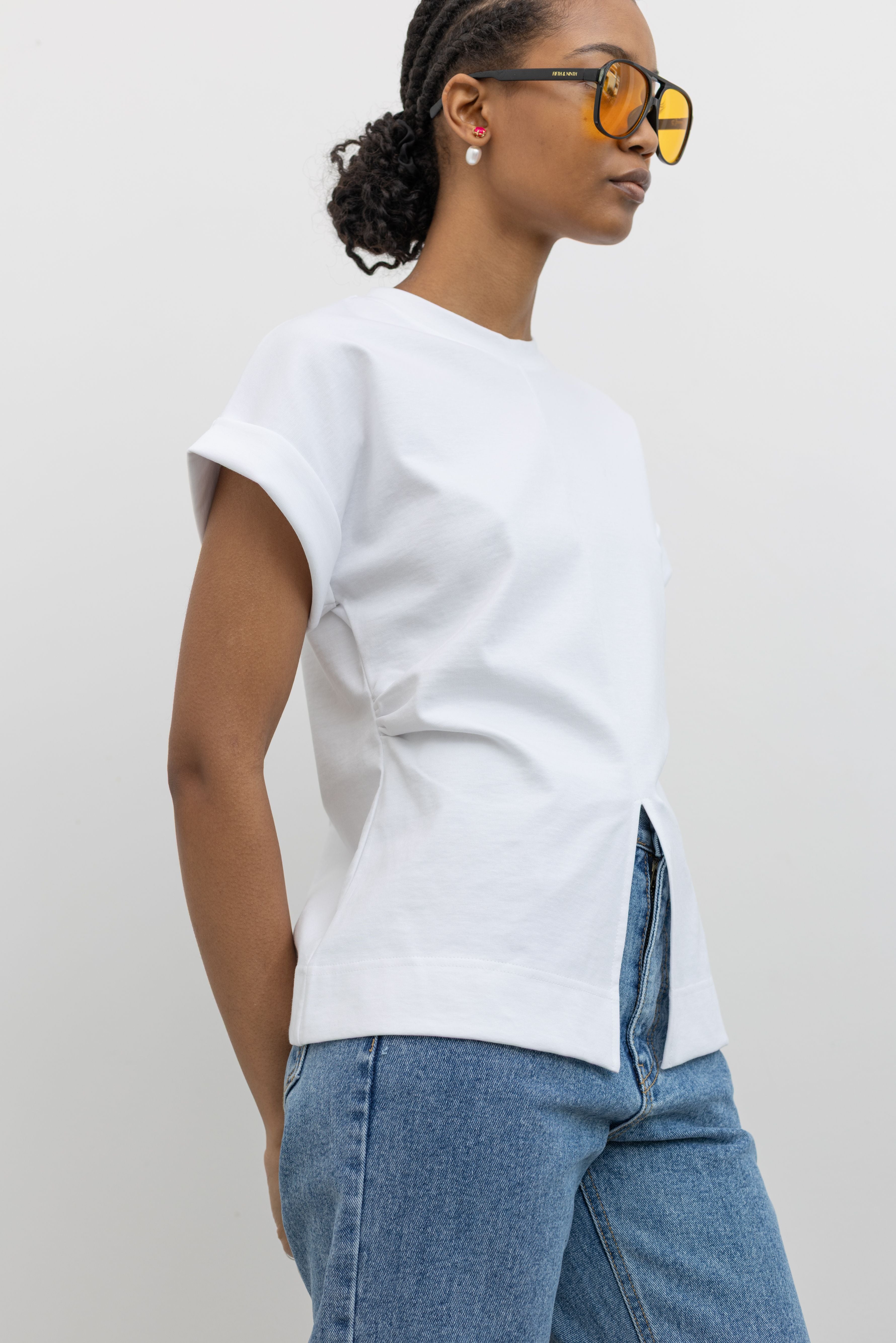 3/4 view of a woman posing with sunglasses wearing an elevated spring staple in a Alas Eius label white cinched waist rounded top designed stitched cuffed short sleeves, ruched side seams and an open v-cut split hem at the front paired with light wash denim jeans at an Alas Eius store. 