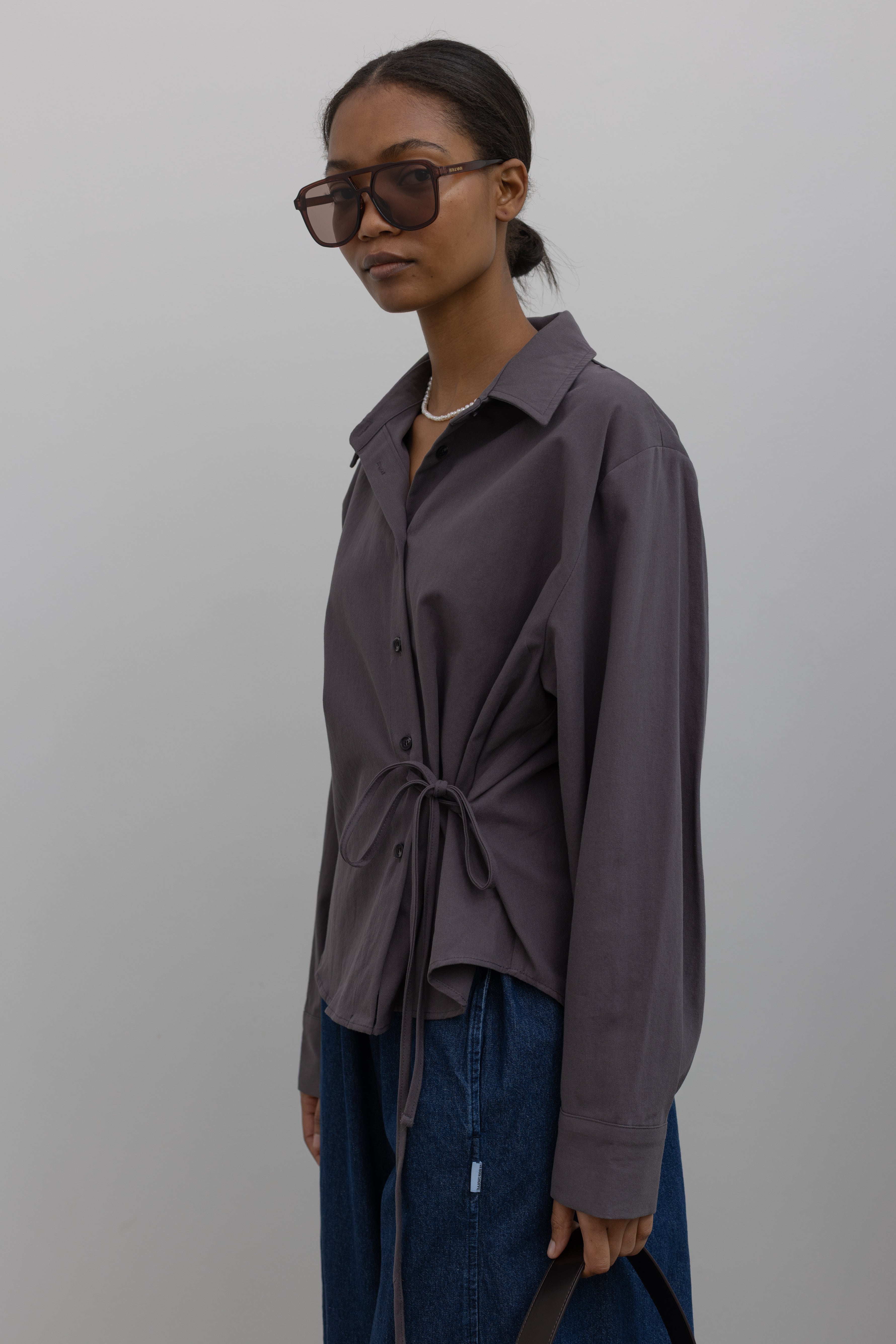 Woman wearing capsule wardrobe essential in an modern and flattering Alas Eius label gray brushed cotton cinched waist button down collared long sleeve top with denim at Alas Eius