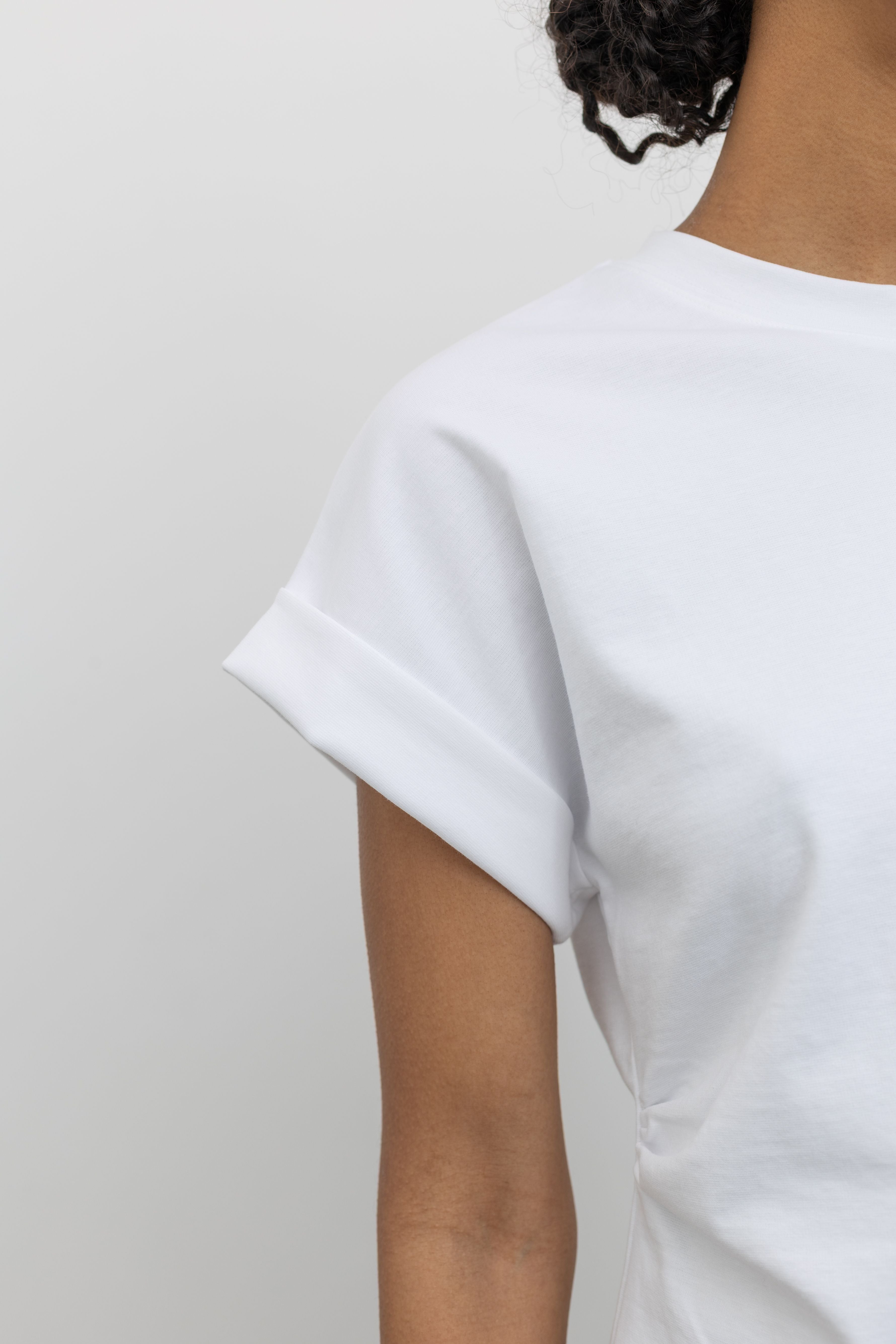 Close-up detailed view of the rolled and stitched short sleeve cuff on the  elevated and chic staple Alas Eius label white cinched waist rounded top featuring an open v-cut vent at an Alas Eius store. 