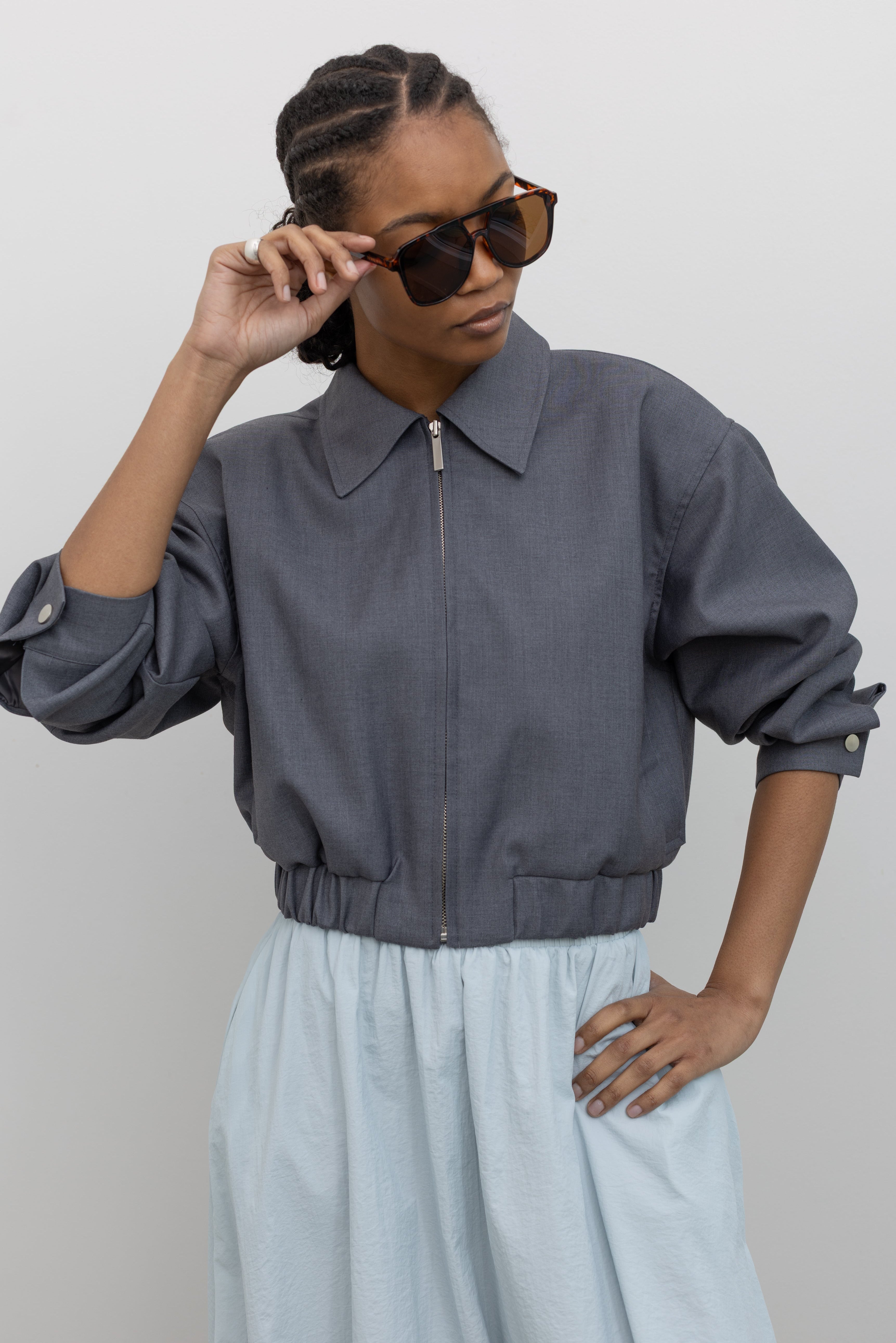Front view of a woman wearing modern minimalist womenswear spring fashion in an Alas Eius label charcoal gray cropped tailored zip jacket featuring an elastic hem for a flattering tailored look at an Alas Eius store. 