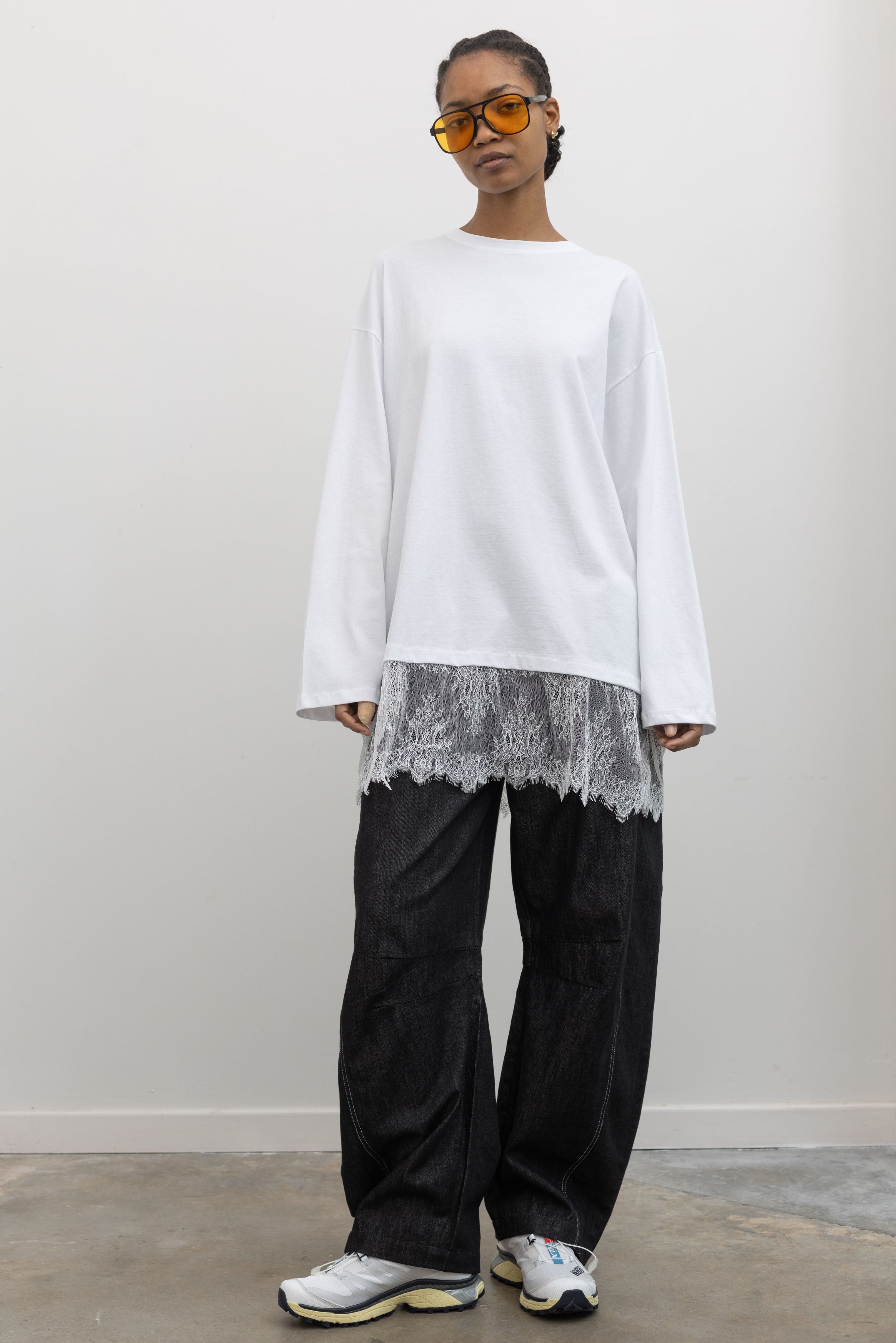 Full length front view of a woman standing wearing spring fashion in a 
versatile Alas Eius label white long-sleeve oversized tunic top with lace hemline paired with Alas Eius dark wash barrel jeans and Salomon sneakers at and Alas Eius store. 