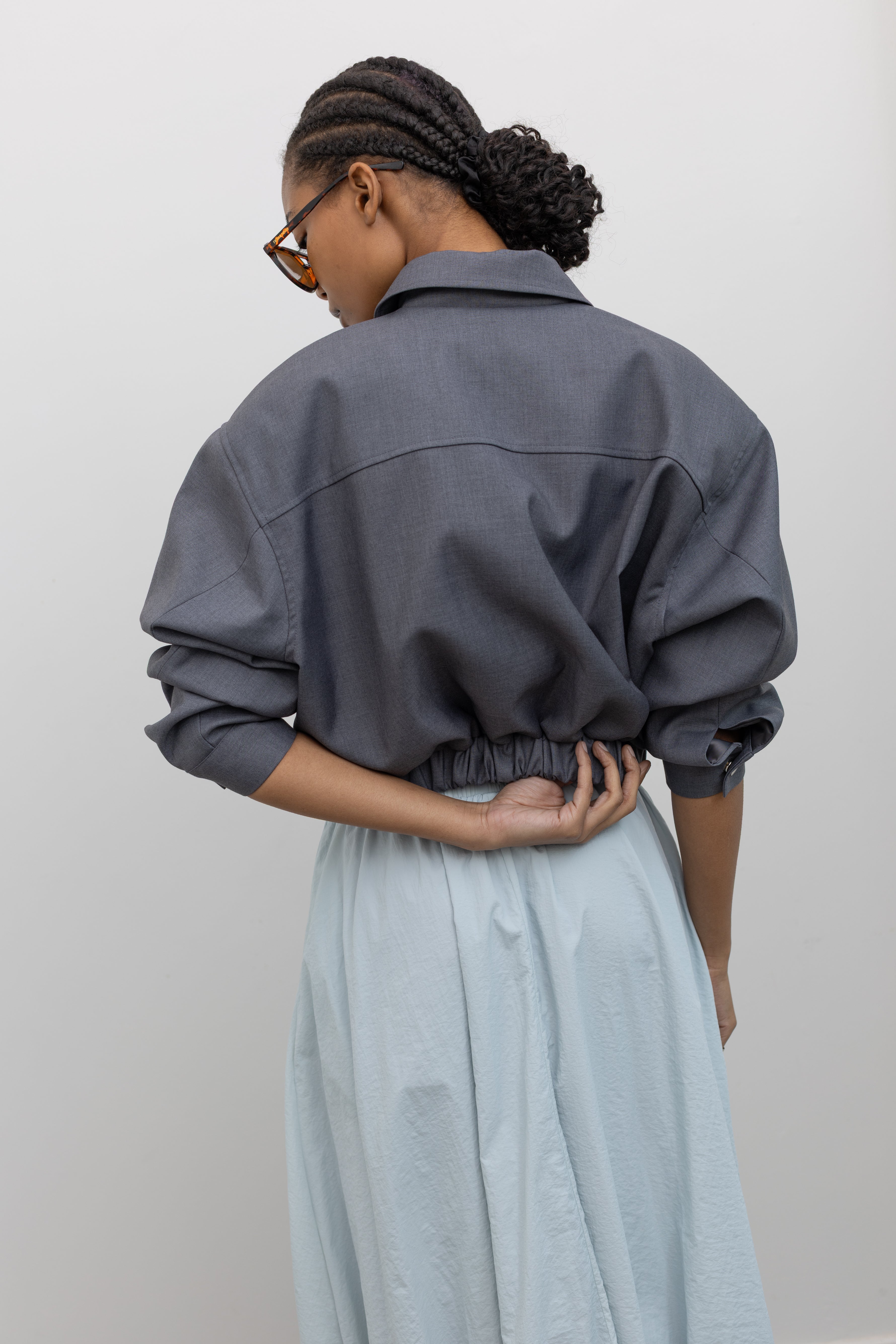 Back view of a woman wearing feminine and chic spring fashion in an Alas Eius label charcoal gray cropped tailored zip jacket featuring an elastic hem for a flattering cinched waist at an Alas Eius store. 