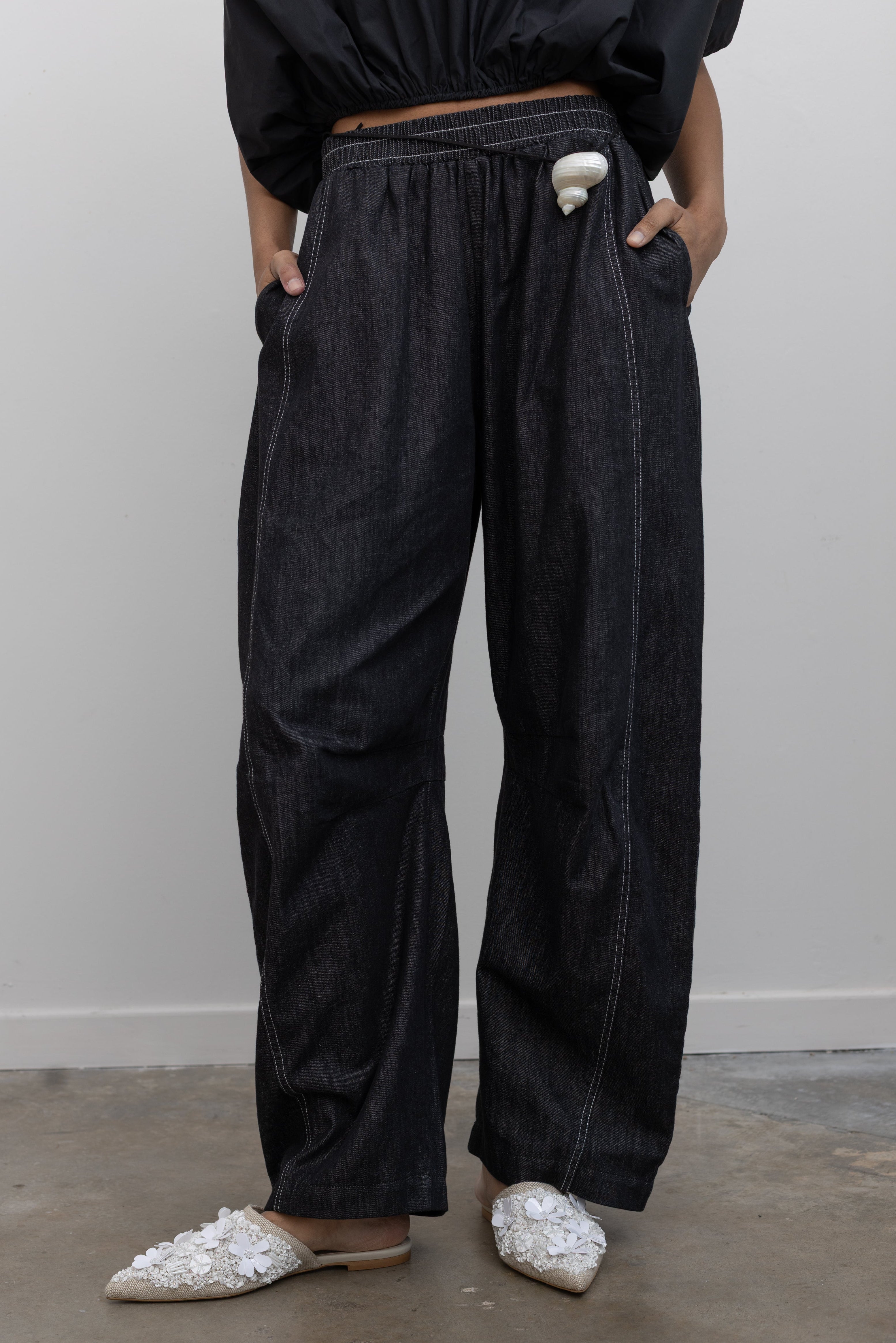 Close-up front view of a woman standing wearing modern minimalist  fashion in the Alas Eius label black cotton barrel leg pants featuring contrasting white double stitching, side seam pockets, and an elastic waistband  at an Alas Eius store. 