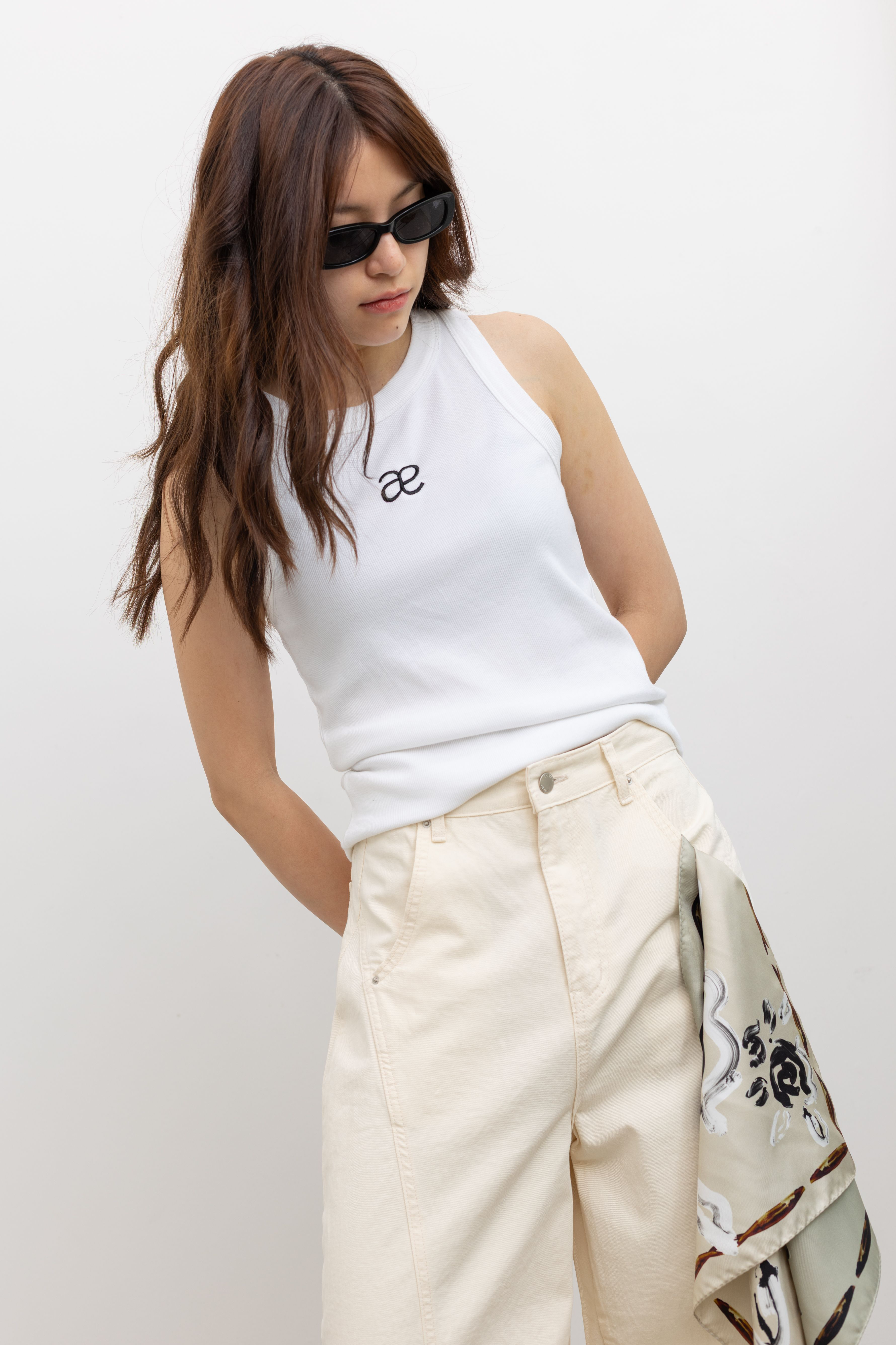 Model wearing an elevated basic in a Alas Eius label white cotton blend ribbed racerback tank top with contrasting embroidered AE logo on chest paired Collective Ivory Balloon Curved Cotton Pant at Alas Eius.